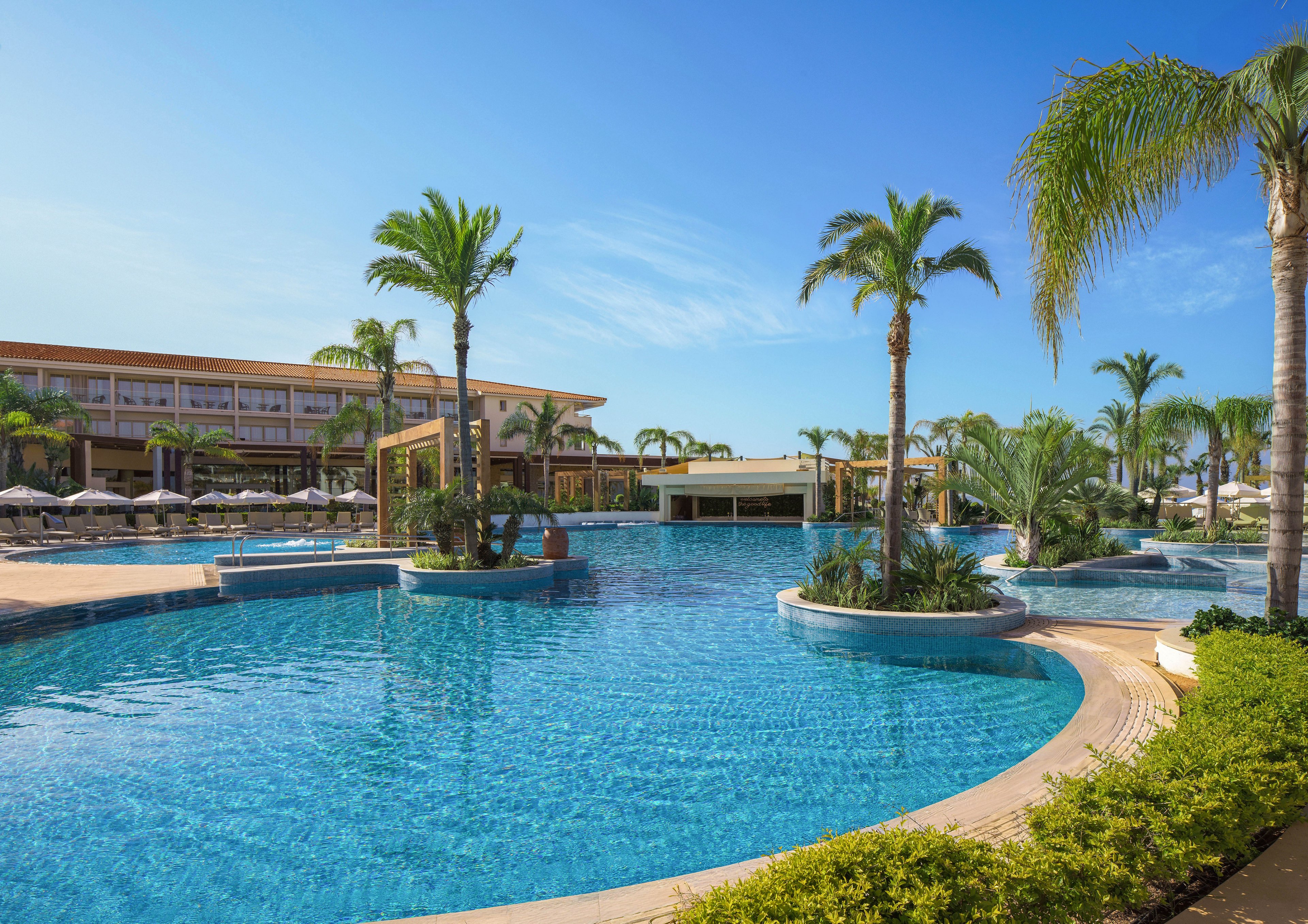 A large, clear blue swimming pool dominates the foreground of a modern resort. Tall palm trees and manicured greenery surround the water, creating a tropical atmosphere. Sun loungers and umbrellas line the far side of the pool in front of a multi-story hotel building. The sky is bright and cloudless, emphasizing a sunny, relaxing vacation setting.