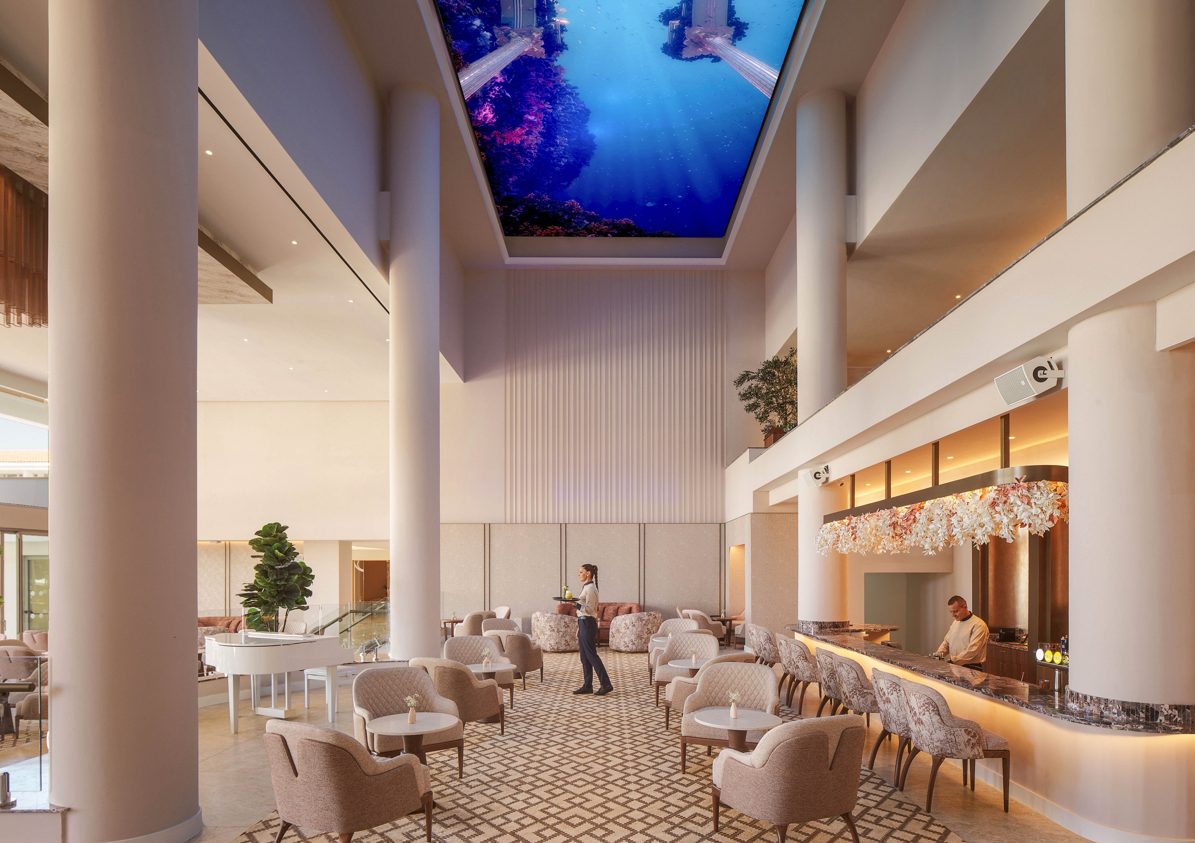 Spacious contemporary hotel lobby features a stylish bar and lounge seating under a dramatic high ceiling. A bartender serves a guest in the center of the room, adding a sense of hospitality and service. Soft neutral furnishings, patterned flooring, and warm lighting create an inviting, upscale atmosphere. A large digital screen on the ceiling displays an underwater scene, adding a unique visual focal point.