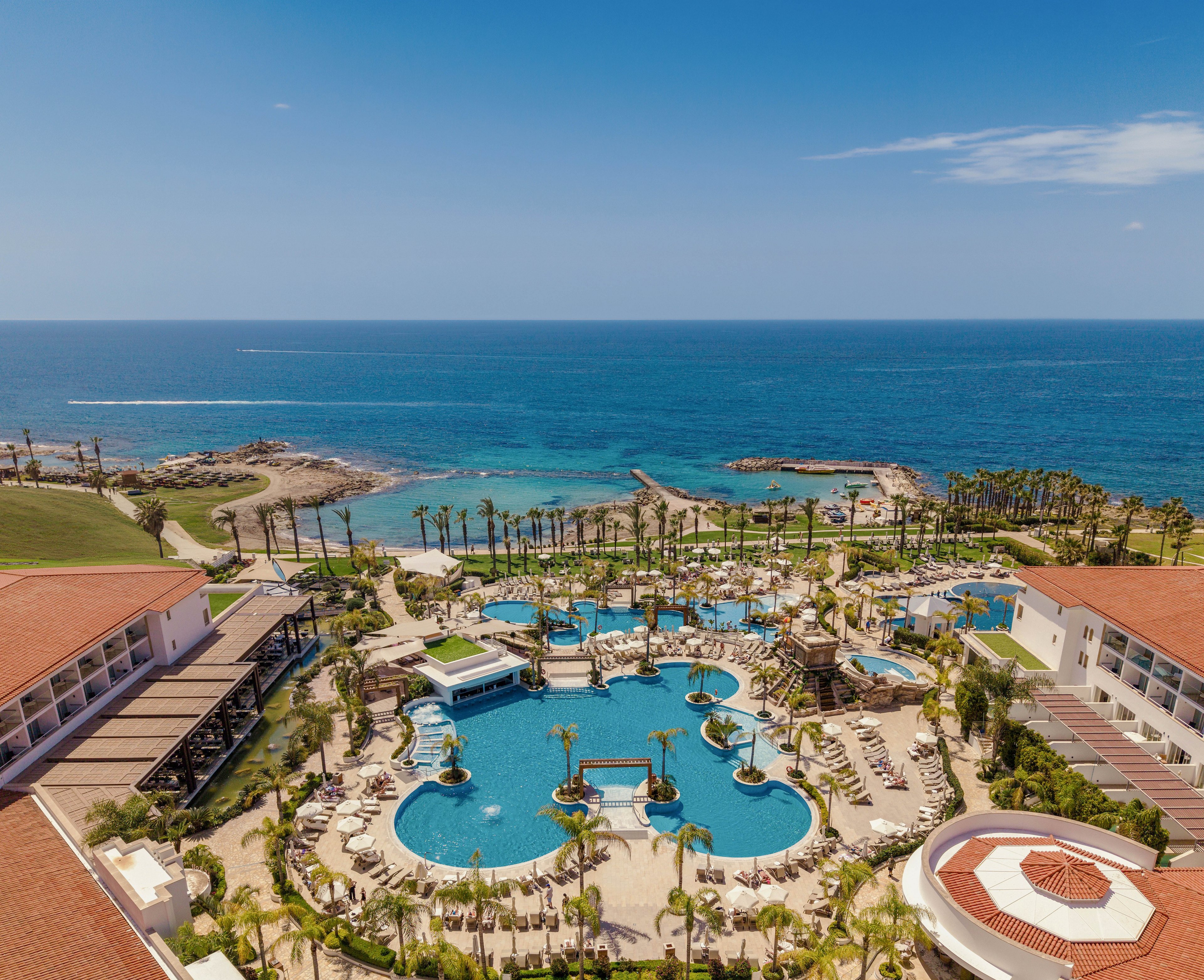 A large luxury resort complex overlooks a bright blue sea on a clear sunny day. Multiple lagoon-style swimming pools with bridges, fountains, and palm trees form the centerpiece of the property. Sun loungers and umbrellas surround the pools, with terracotta-roofed buildings framing the scene. The coastline, rocky shore, and distant horizon emphasize the tropical vacation setting.
