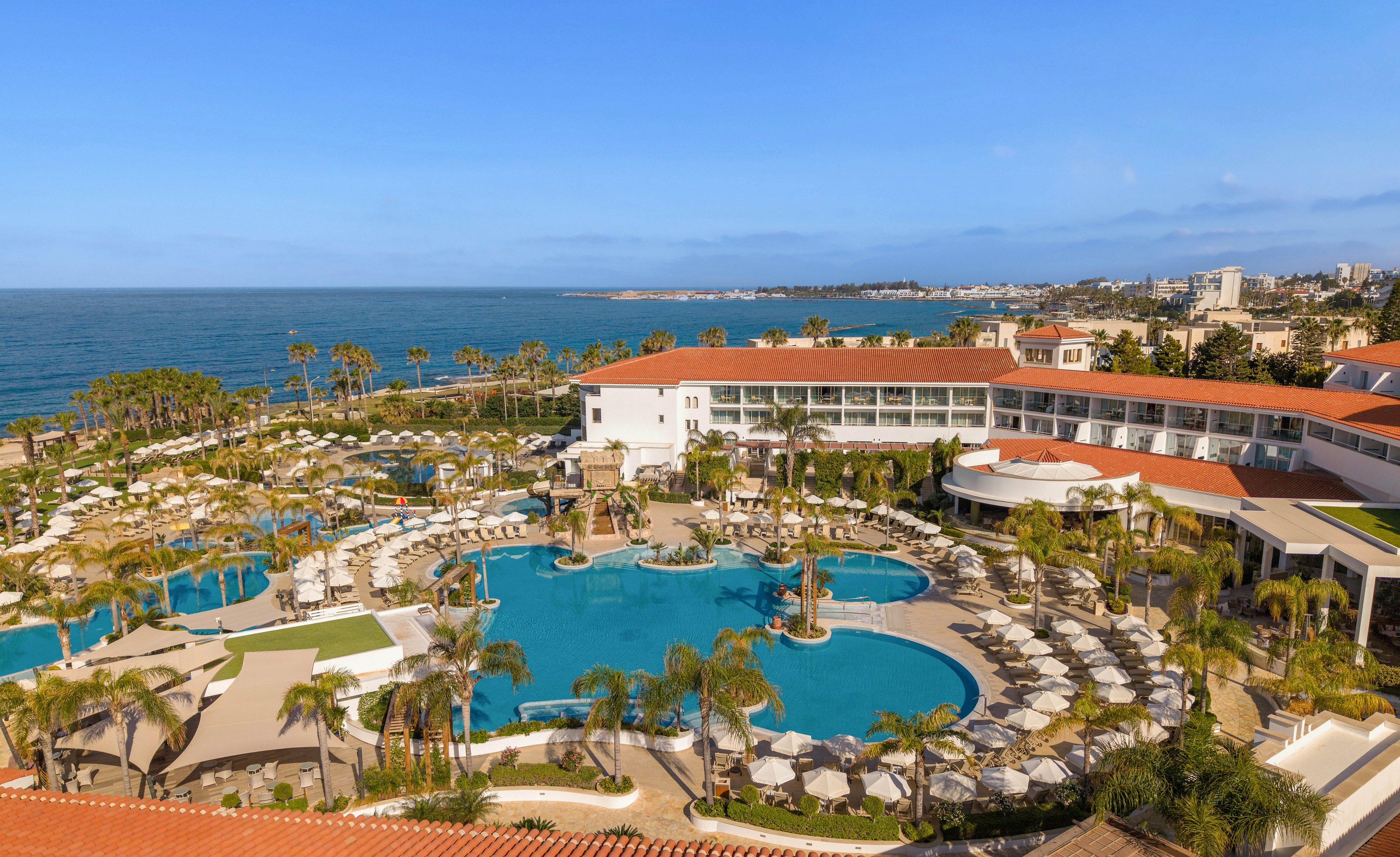 A large seaside resort features multiple outdoor swimming pools surrounded by palm trees and sun loungers. The white and terracotta-roofed buildings overlook the coastline and open sea under a clear blue sky. Guests relax around the curving pools, creating a tranquil vacation atmosphere. The scene suggests a warm, sunny day at a coastal holiday destination.