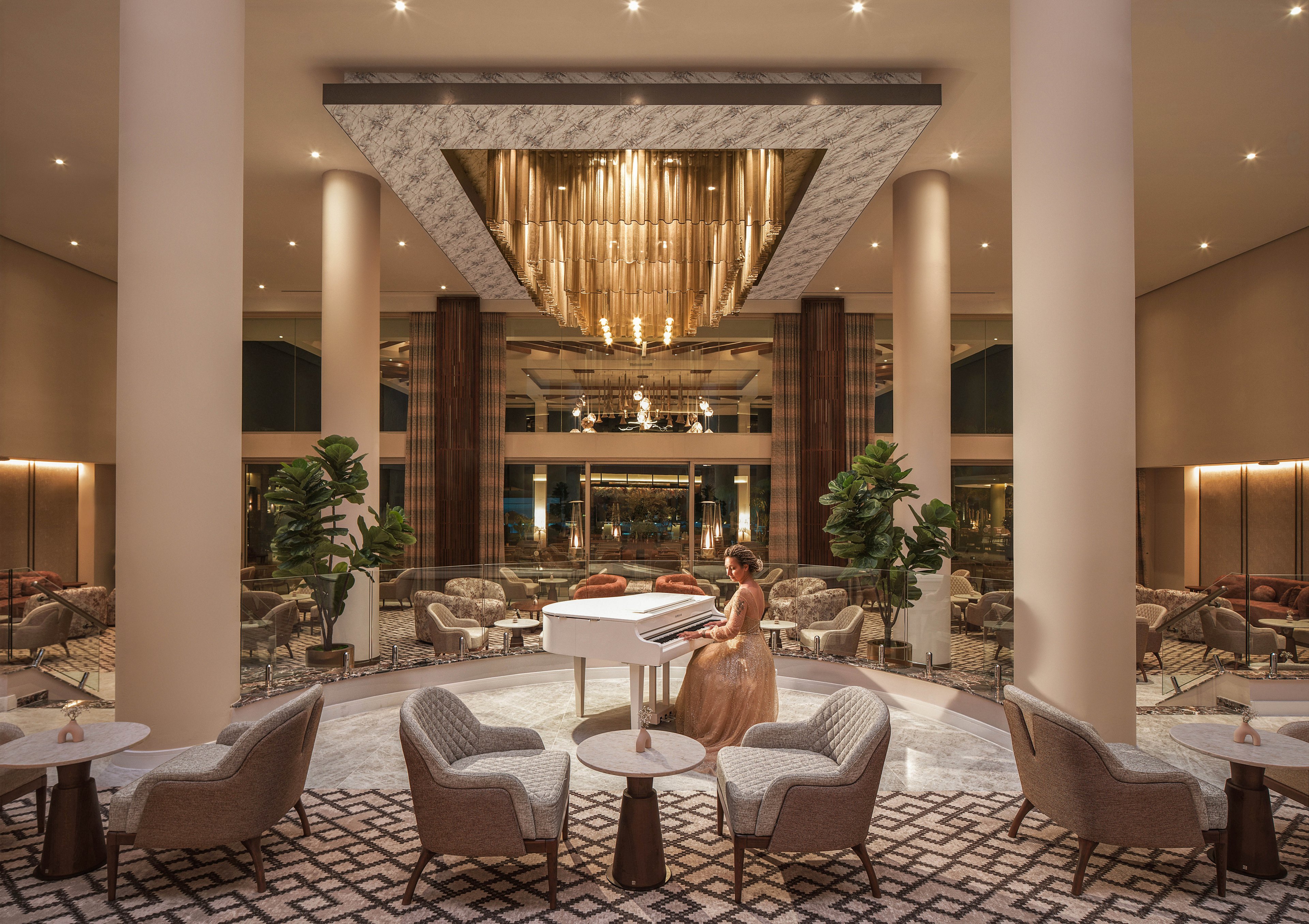 A woman in an evening gown plays a white grand piano in a luxurious hotel lounge. The spacious interior features tall columns, plush armchairs, and small round tables arranged on a patterned carpet. Warm ambient lighting and a dramatic ceiling fixture create a sophisticated atmosphere. Large windows and decorative plants add depth and a sense of openness to the refined setting.