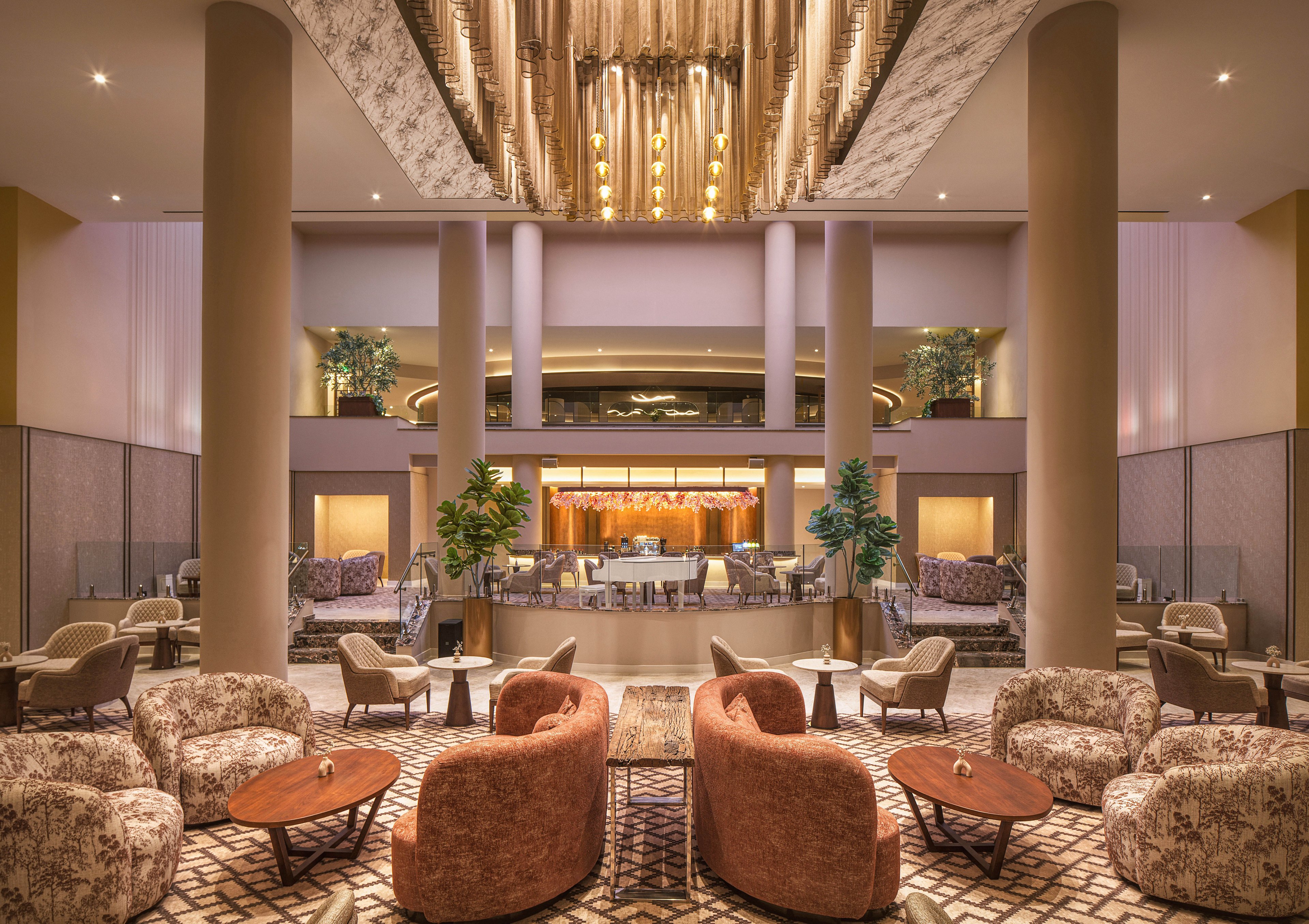 Spacious hotel lobby lounge featuring plush armchairs and patterned sofas arranged around low wooden tables. Tall columns frame the central bar area in the background, highlighted by warm ambient lighting. A dramatic chandelier hangs from the ceiling, adding elegance to the contemporary interior. Potted plants and soft neutral tones create a welcoming, upscale atmosphere suitable for socializing or relaxing.