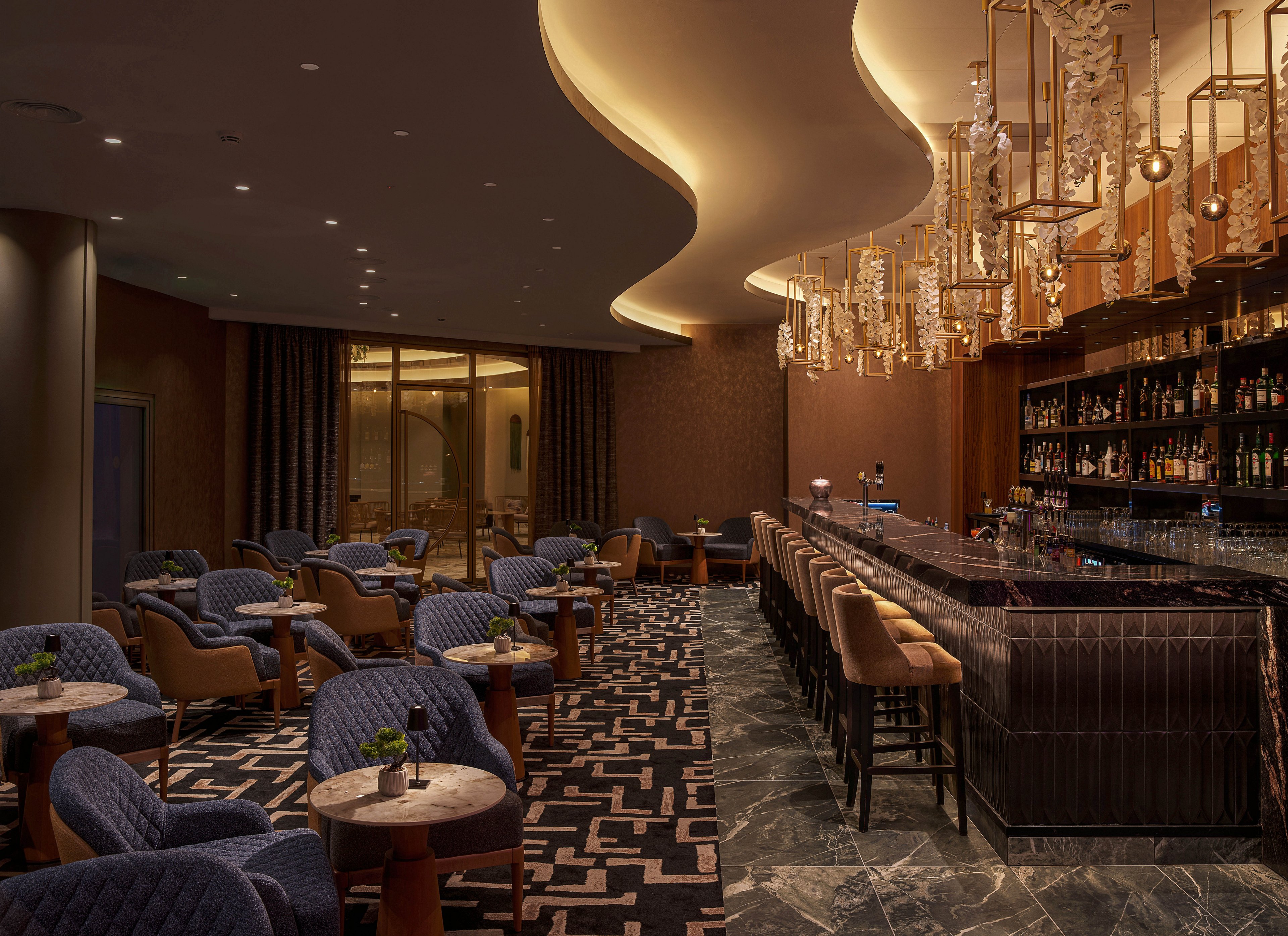 Upscale lounge bar interior featuring a long marble-topped counter lined with upholstered bar stools. Plush blue armchairs surround small round tables with simple green floral centerpieces. Warm ambient lighting, sculptural ceiling details, and cascading chandeliers create an elegant atmosphere. A well-stocked back bar with bottles and glassware adds to the sophisticated nighttime setting.