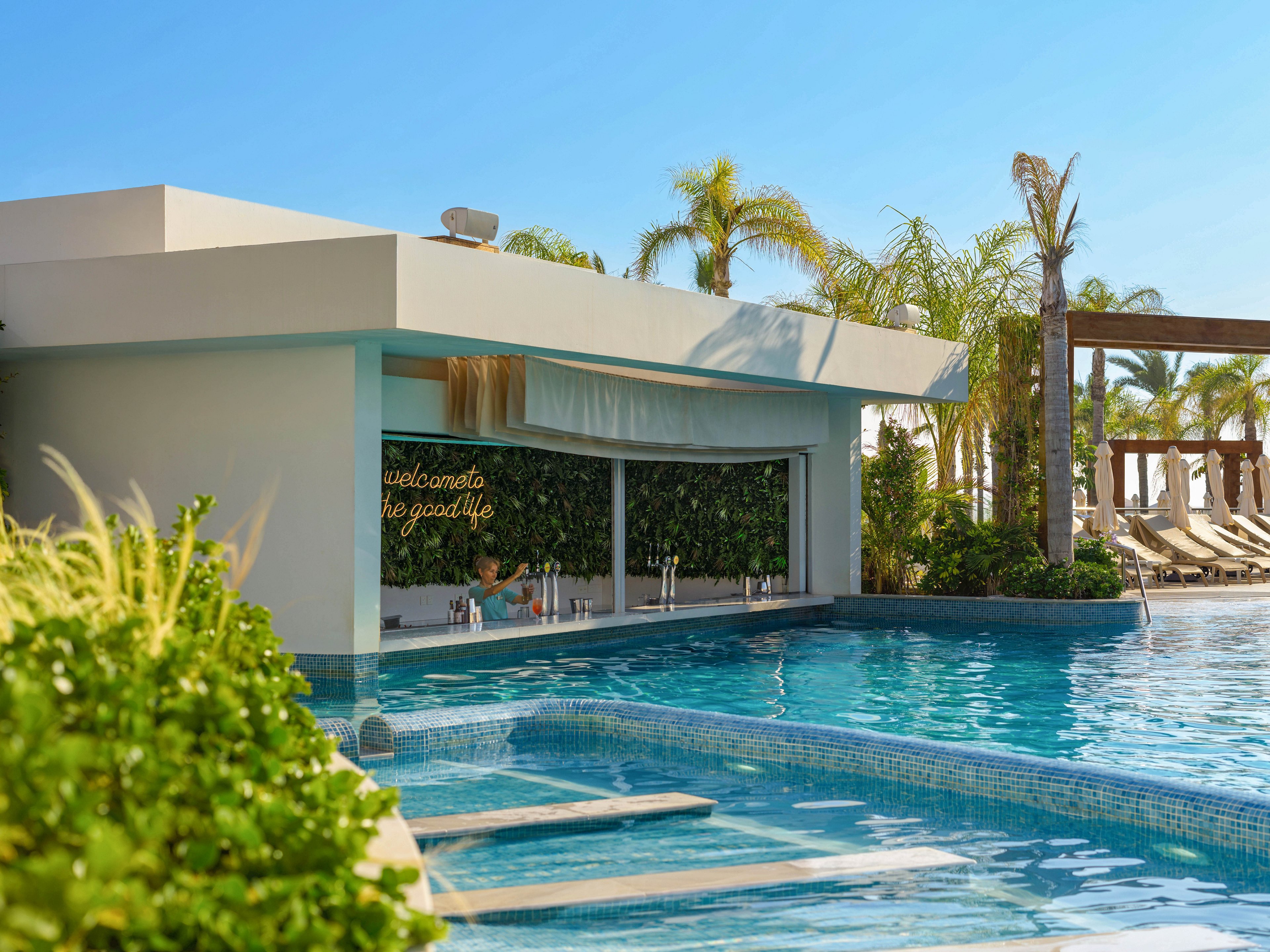A modern resort pool features a swim-up bar framed by lush greenery and palm trees. Lounge chairs line the deck in the background under clear blue skies. The water is calm and inviting, reflecting the contemporary architecture of the bar structure. A neon-style sign inside the bar area reads "welcome to the good life," enhancing the relaxed vacation atmosphere.