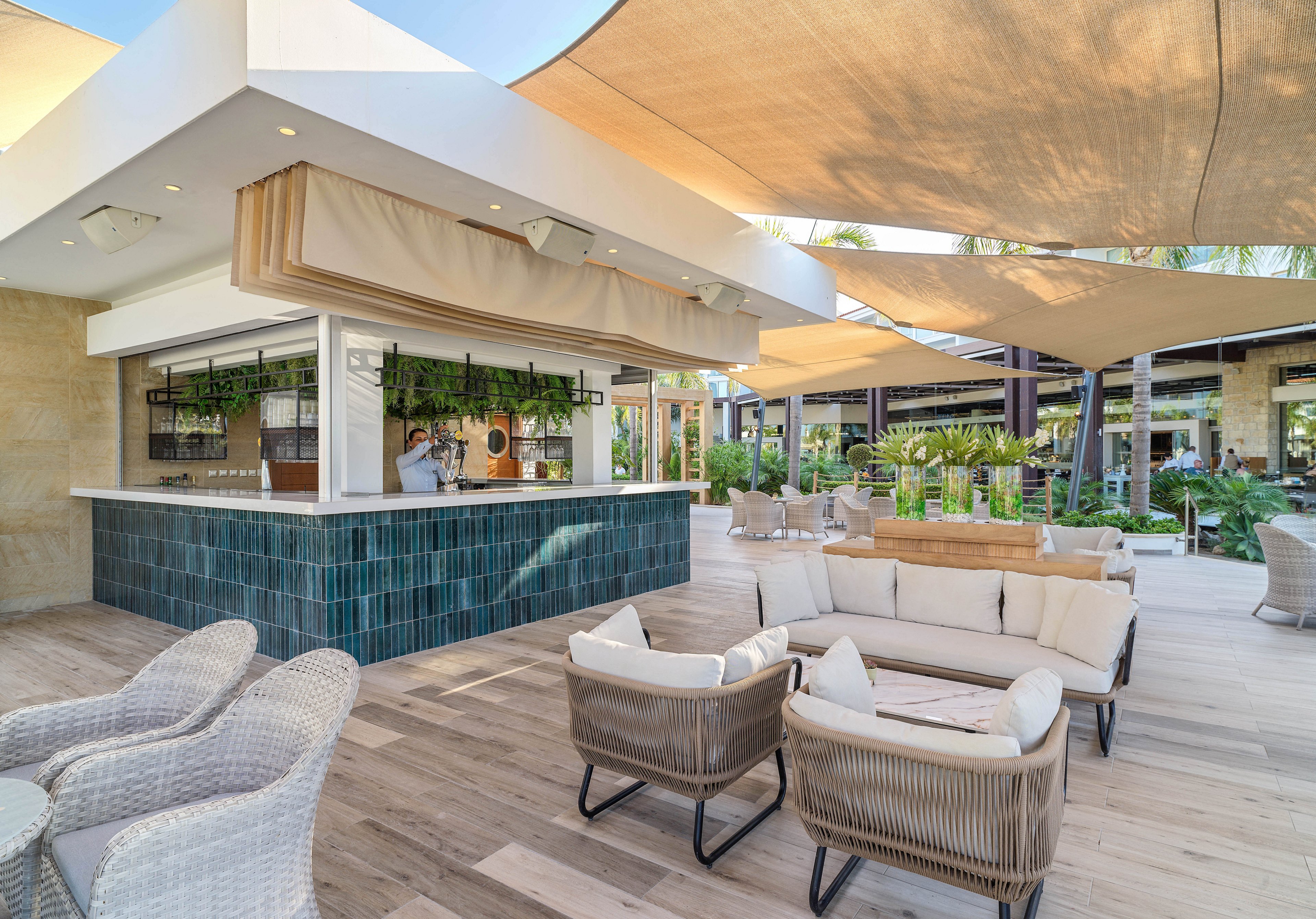 An open-air bar sits at the center of a stylish outdoor lounge area. Comfortable wicker chairs and cushioned sofas surround low tables on a light wooden deck. Large fabric shade sails provide cover from the sun while greenery and potted plants add a tropical feel. The setting appears relaxed and inviting, ideal for socializing or enjoying drinks in a resort environment.