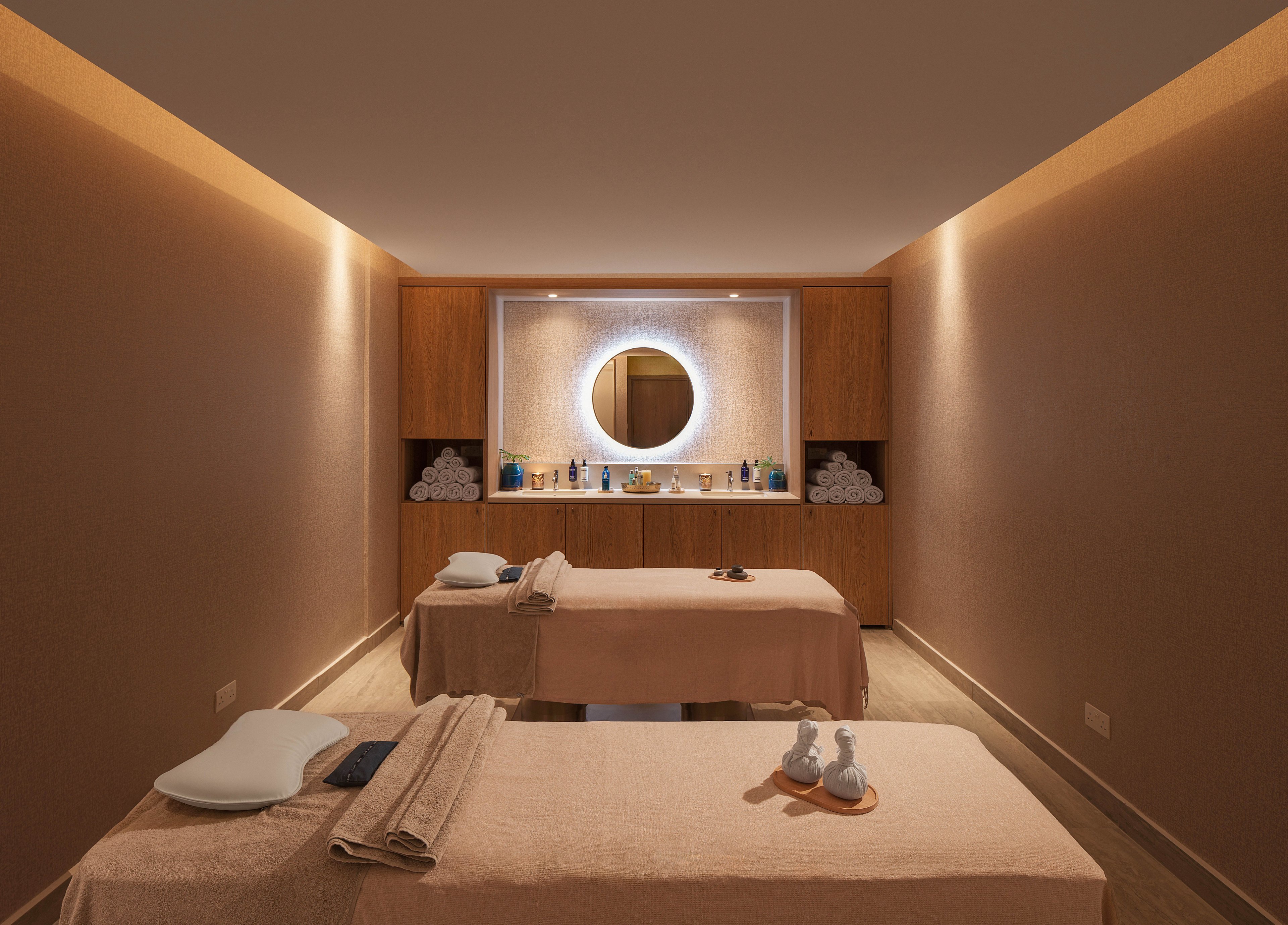 A softly lit spa treatment room features two massage tables arranged side by side. Neutral beige tones and wooden cabinetry create a warm, calming atmosphere. Rolled white towels and various bottles of oils and lotions are neatly displayed on shelves. A round illuminated mirror on the back wall adds a modern touch to the serene wellness space.