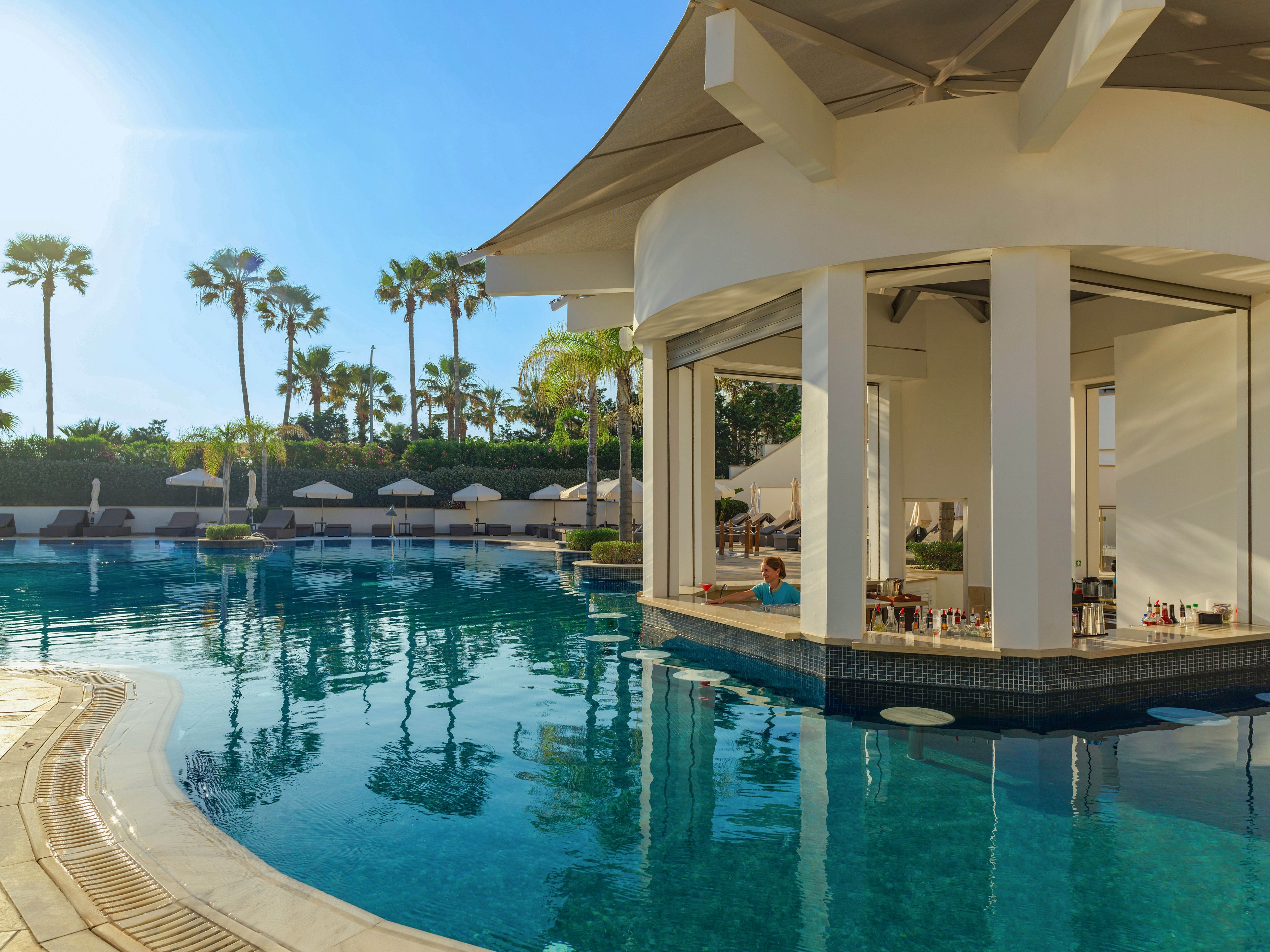 A luxurious resort swimming pool features a circular swim-up bar surrounded by clear blue water. A guest sits at the bar while drinks and bottles are displayed on the counter inside the open pavilion. Palm trees and rows of white sun loungers with umbrellas line the background, suggesting a tropical vacation setting. Warm sunlight reflects off the water, creating a relaxed and inviting atmosphere for leisure and refreshment.