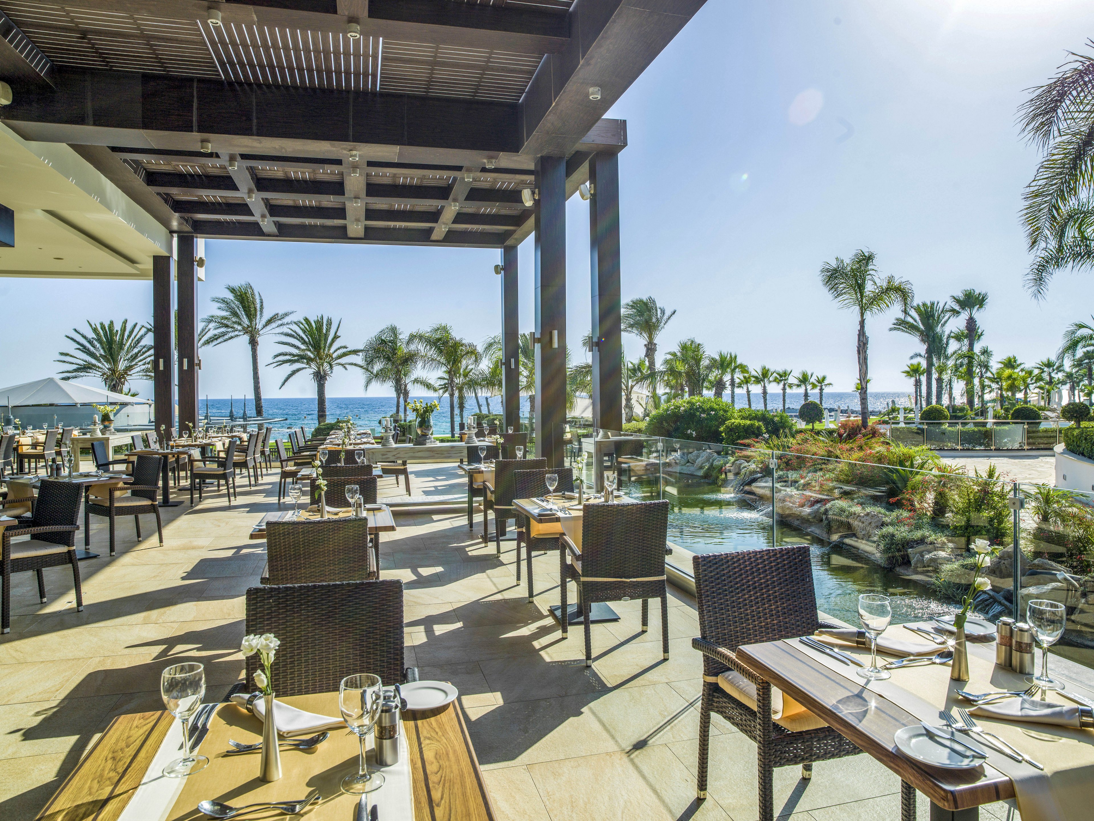 An open-air restaurant terrace overlooks a lush garden and the sea beyond. Wooden tables are neatly set with plates, cutlery, wine glasses, and napkins, ready for guests. Palm trees and tropical plants surround the outdoor dining area, creating a resort-like atmosphere. Bright sunlight and clear blue sky give the scene a relaxed, vacation mood.