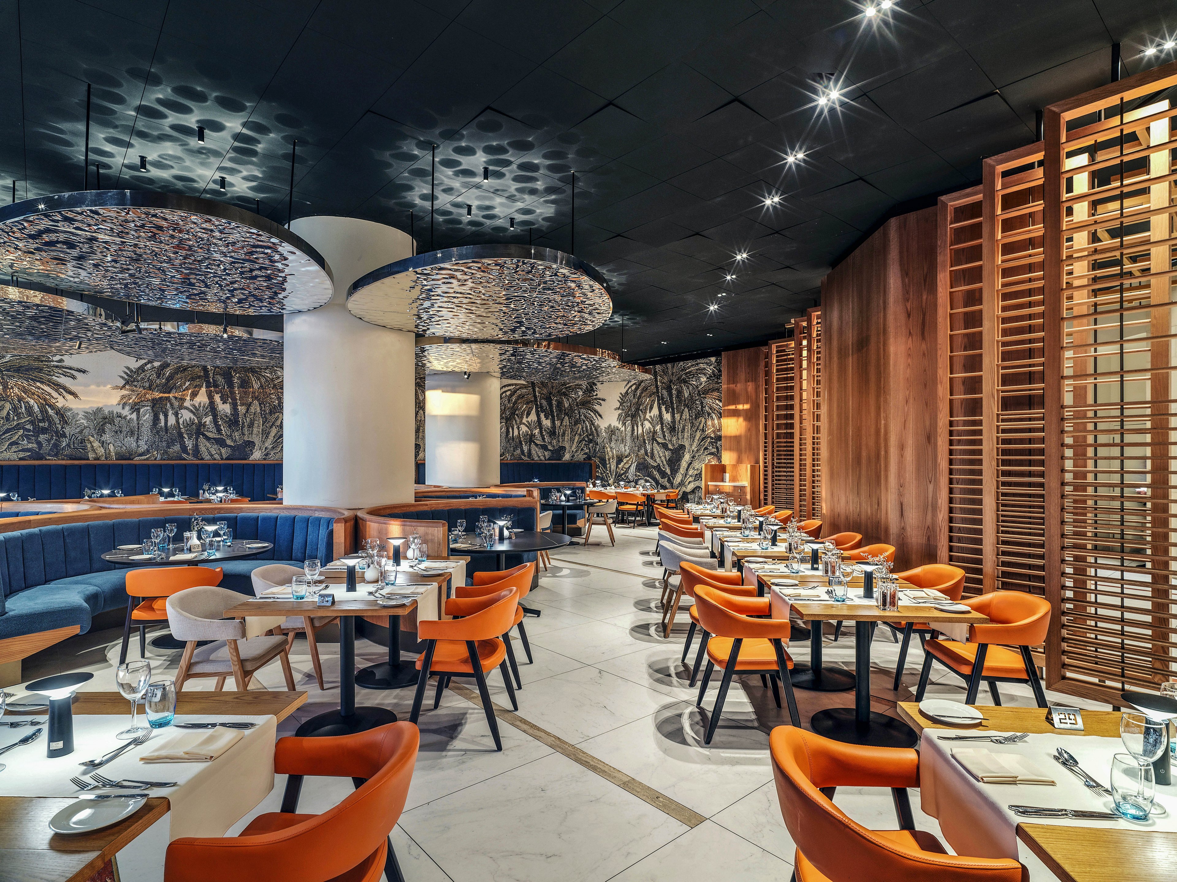 Contemporary restaurant dining room featuring neatly set tables and stylish seating. The space includes orange and gray chairs, curved banquettes, and wooden accents that create a warm, inviting atmosphere. Large circular ceiling fixtures and a monochrome wall mural add visual interest and sophistication. The restaurant appears ready for guests, with glassware, cutlery, and menus arranged on each table.