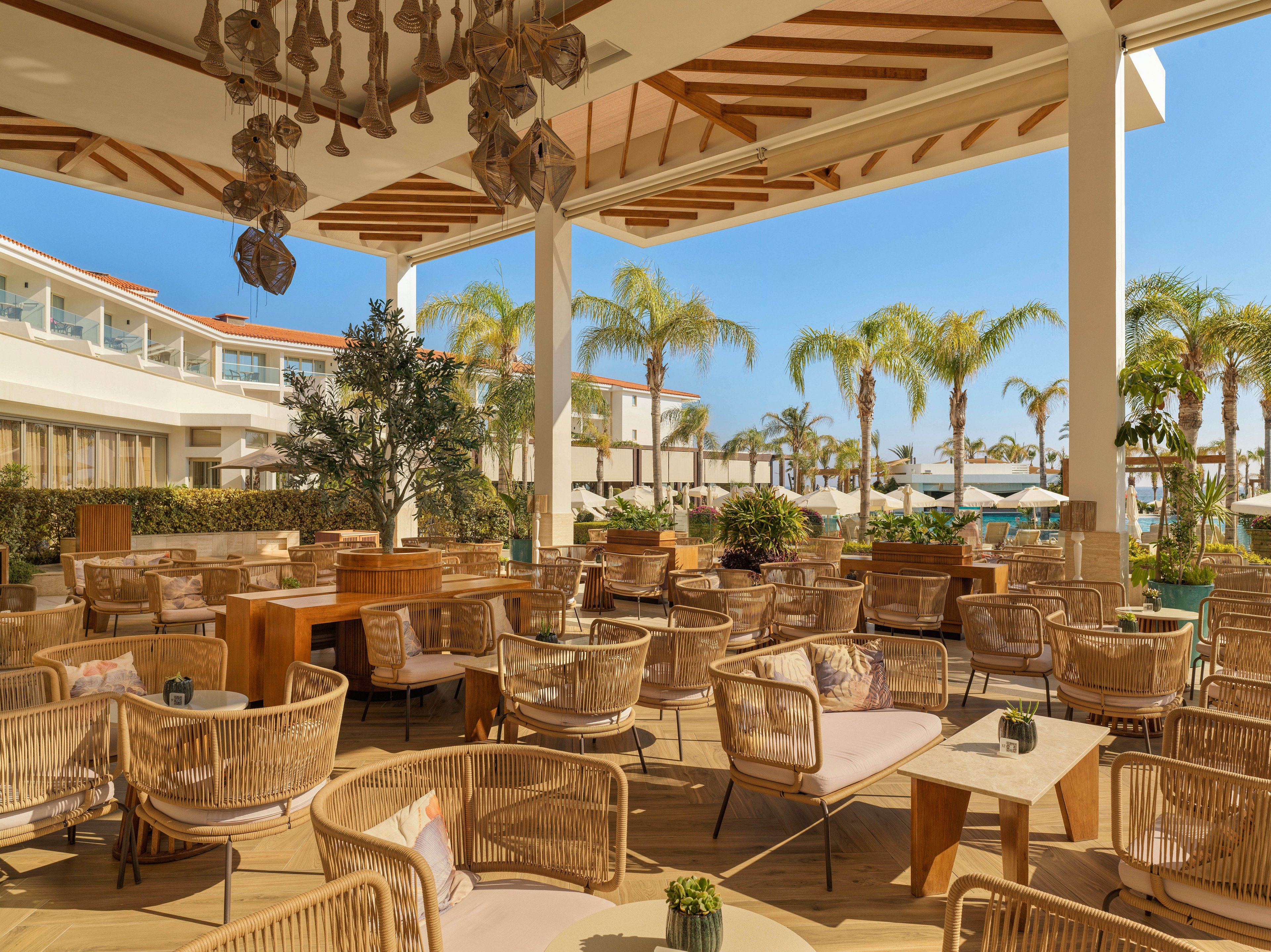 An open-air resort lounge features numerous wicker chairs and low tables arranged on a wooden deck. The setting overlooks a pool area with sun loungers and umbrellas under a clear blue sky. Palm trees and manicured greenery create a tropical atmosphere around the seating area. Warm neutral tones, natural materials, and soft cushions contribute to a relaxed, upscale vacation mood.