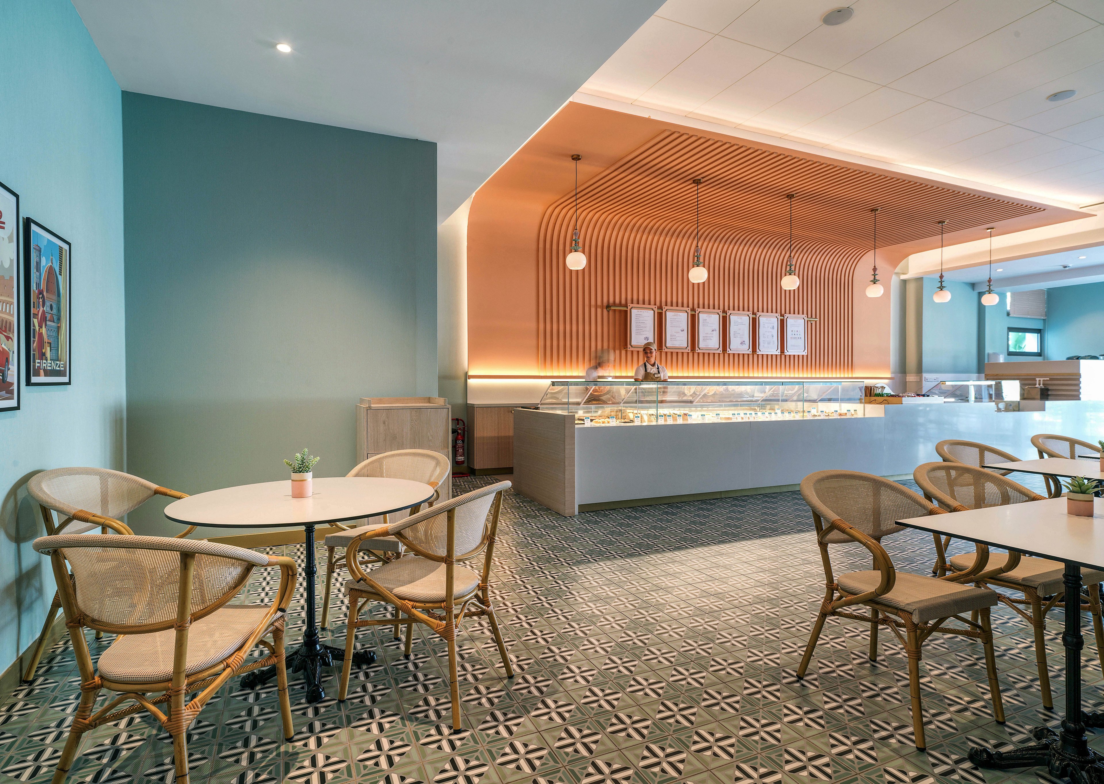 Contemporary cafe interior featuring a sleek service counter and patterned tile floor. Light wooden chairs and round tables are arranged neatly in the foreground, creating an inviting seating area. The back wall showcases a curved wood slat design, hanging pendant lights, and a menu board above the counter. Soft teal walls and warm lighting give the space a calm, modern atmosphere.