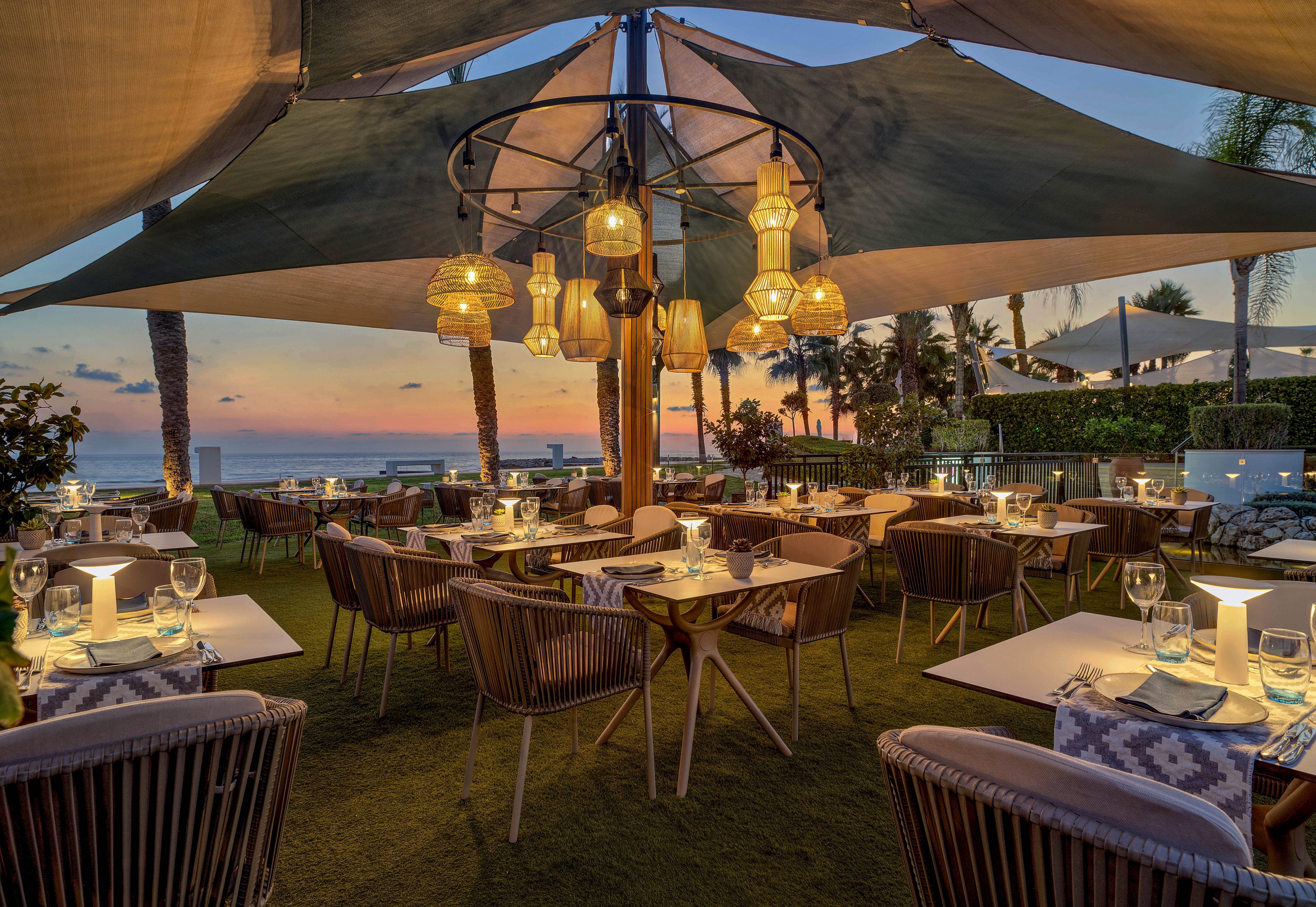 An elegant outdoor restaurant is set for evening dining under large shade sails and hanging lanterns. Tables are neatly arranged with white tablecloths, glassware, and glowing candles, creating an inviting atmosphere. The scene overlooks the sea at sunset, with palm trees framing the view and warm colors in the sky. Soft lighting and comfortable woven chairs contribute to a relaxed yet upscale coastal ambiance.