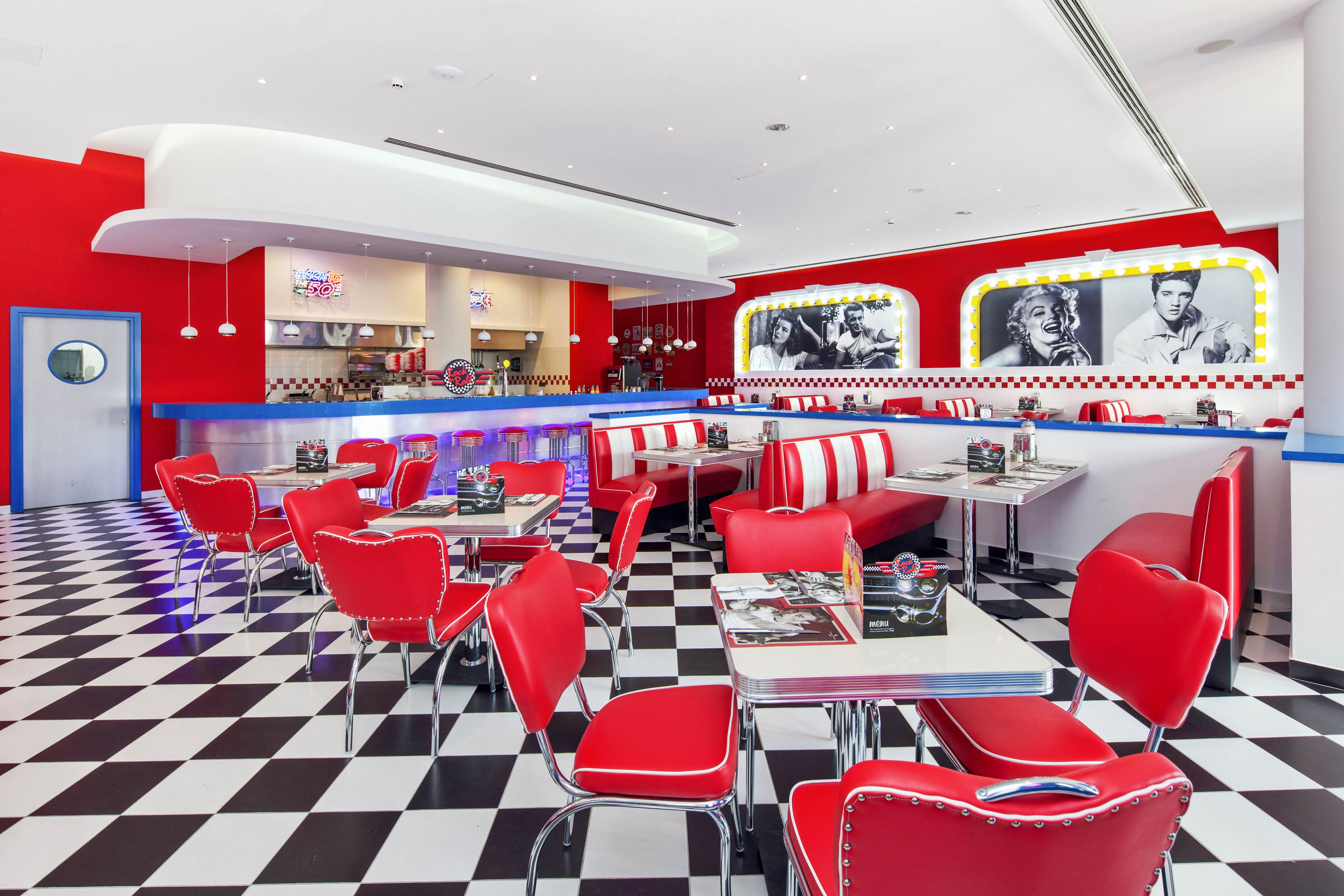 A brightly lit retro American diner features red vinyl booths and chrome-trimmed chairs arranged on a black and white checkered floor. The interior includes a long counter with bar stools and an open kitchen area in the background. Walls are painted in bold red and white, with large black-and-white celebrity photos adding to the nostalgic theme. The overall atmosphere is clean, vibrant, and reminiscent of classic 1950s diners.