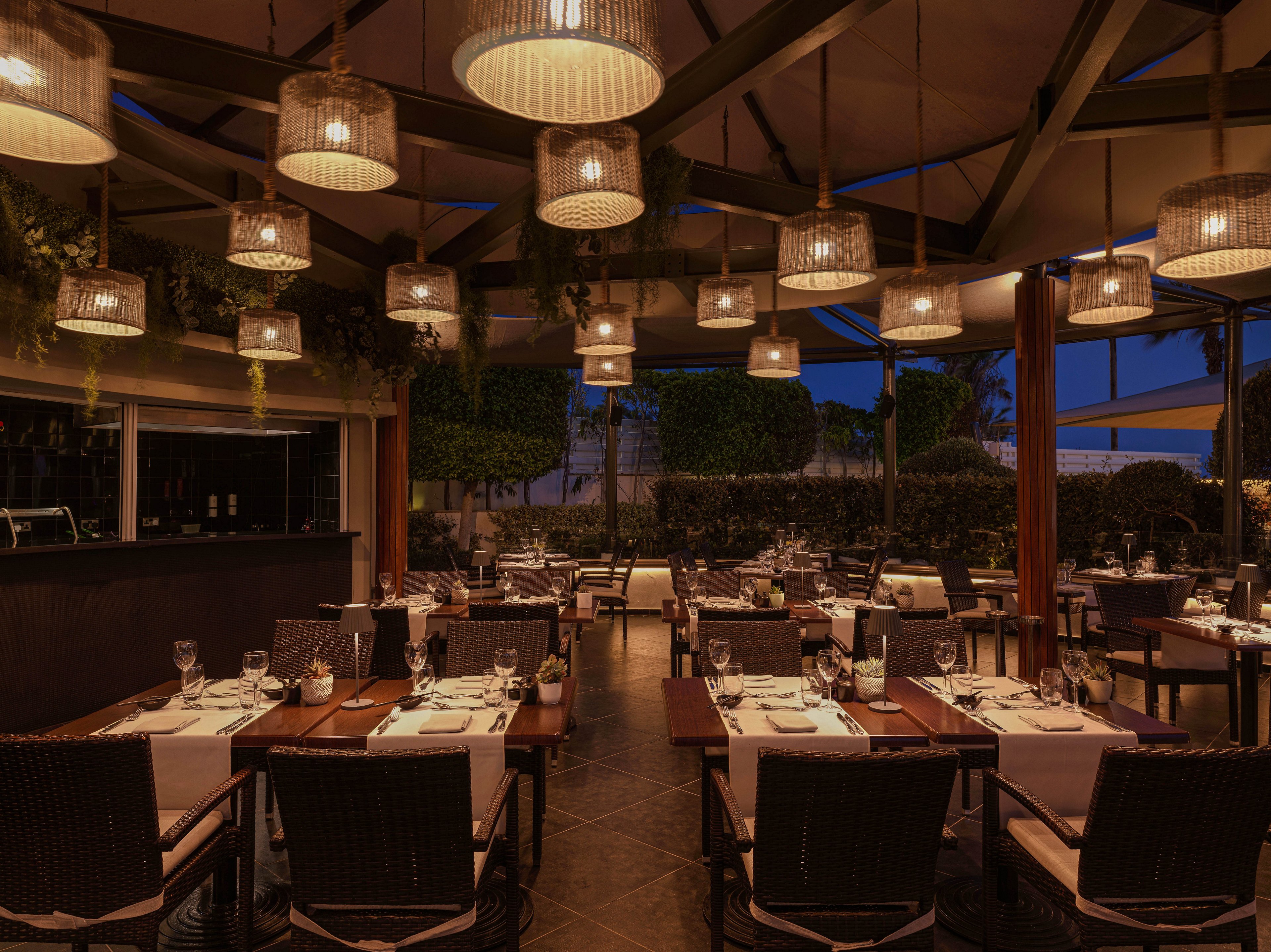 An upscale open-air restaurant is set for evening service under a canopy at dusk. Tables are neatly arranged with white tablecloths, glassware, and folded napkins, creating a refined dining atmosphere. Warm pendant lights hang from the ceiling structure, casting a soft glow over the seating area. Lush greenery and manicured hedges surround the space, adding a sense of privacy and sophistication.