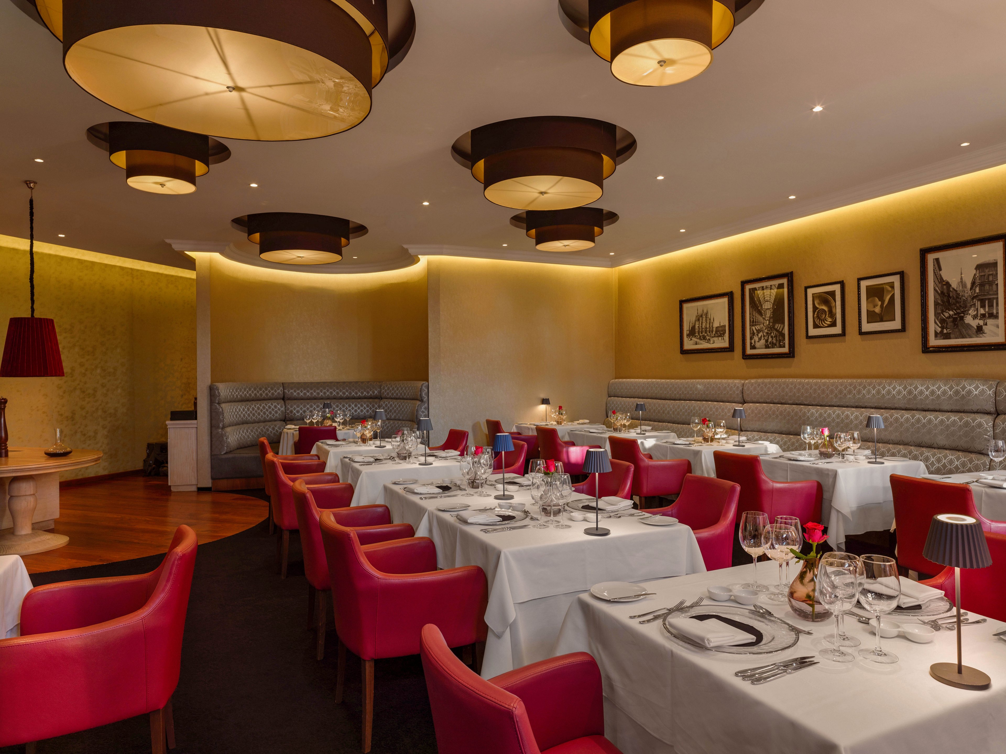 Upscale restaurant interior featuring neatly set tables with white tablecloths and glassware. Red upholstered chairs contrast with gray banquette seating along the wall. Warm ambient lighting from modern circular ceiling fixtures creates a cozy atmosphere. Framed black-and-white pictures decorate the walls, enhancing the sophisticated dining setting.