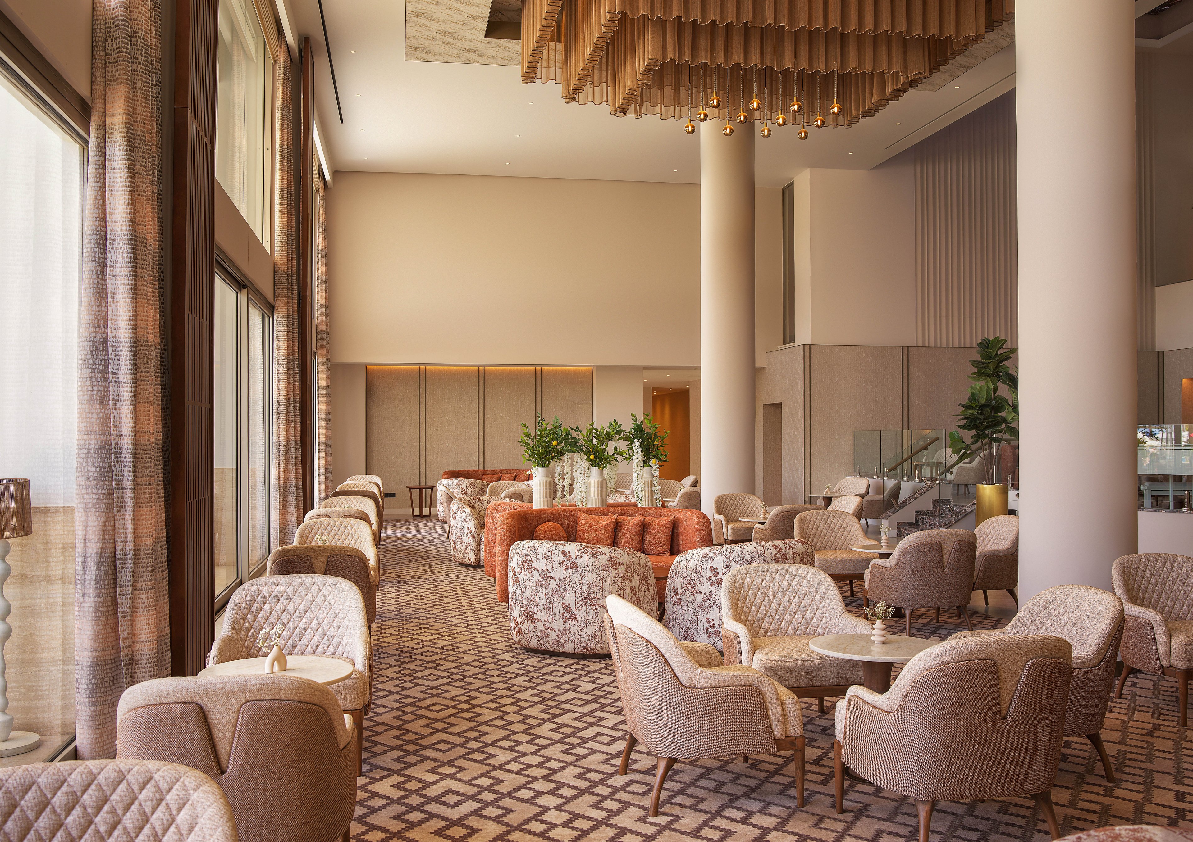 Spacious hotel lounge featuring plush armchairs and round tables arranged in conversational groupings. Large floor-to-ceiling windows on one side allow natural light to fill the room. Warm neutral tones, patterned carpet, and soft fabric textures create a sophisticated atmosphere. Decorative ceiling lights and indoor plants add to the upscale, relaxing setting.