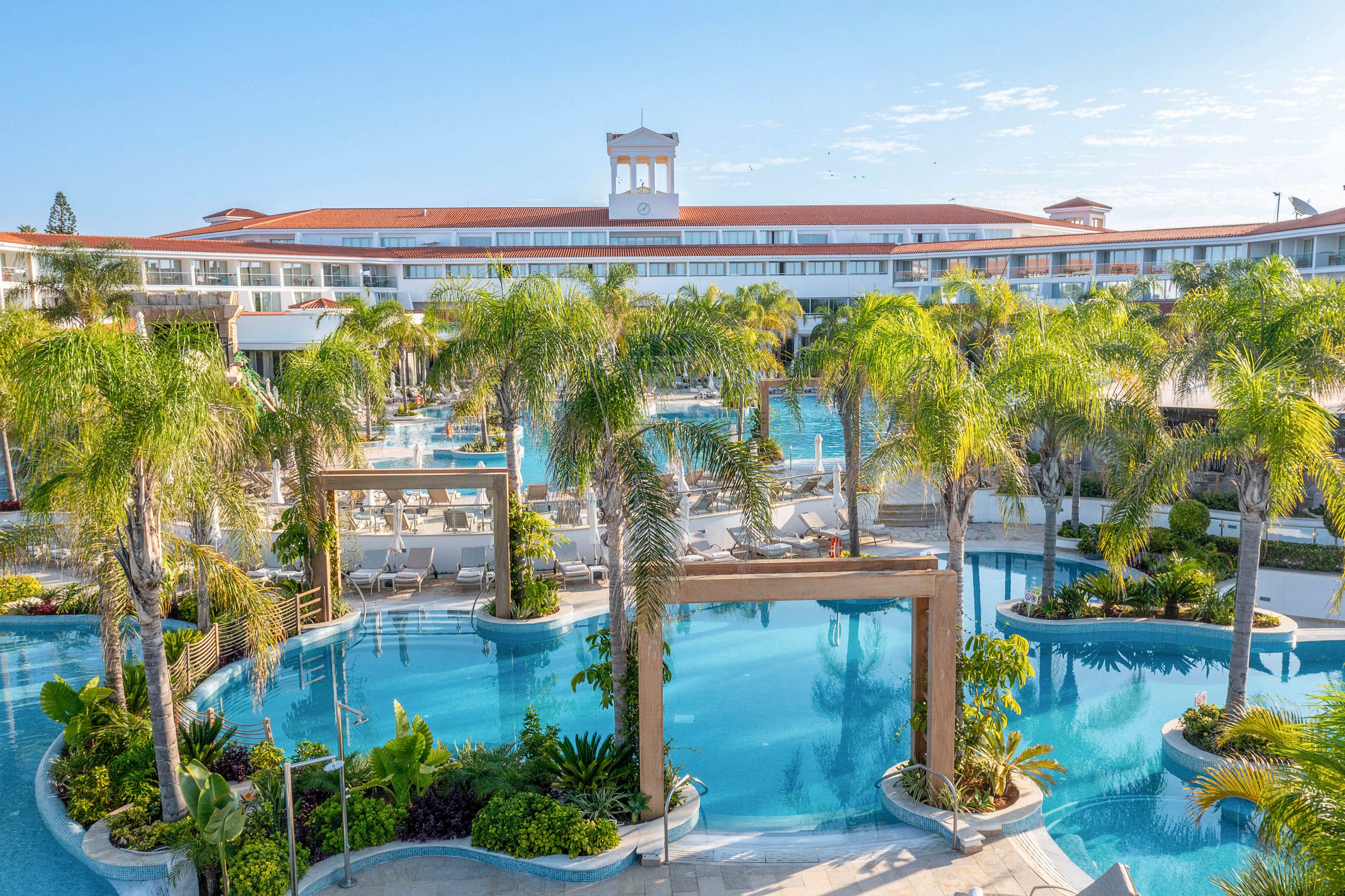 A large resort complex features an expansive outdoor swimming pool surrounded by lush palm trees. Multiple interconnected pools curve around small islands of greenery, creating a tropical oasis atmosphere. Sun loungers and shaded cabanas line the water’s edge, suggesting a relaxing vacation setting. The bright blue sky and calm water emphasize a warm, sunny day at the resort.