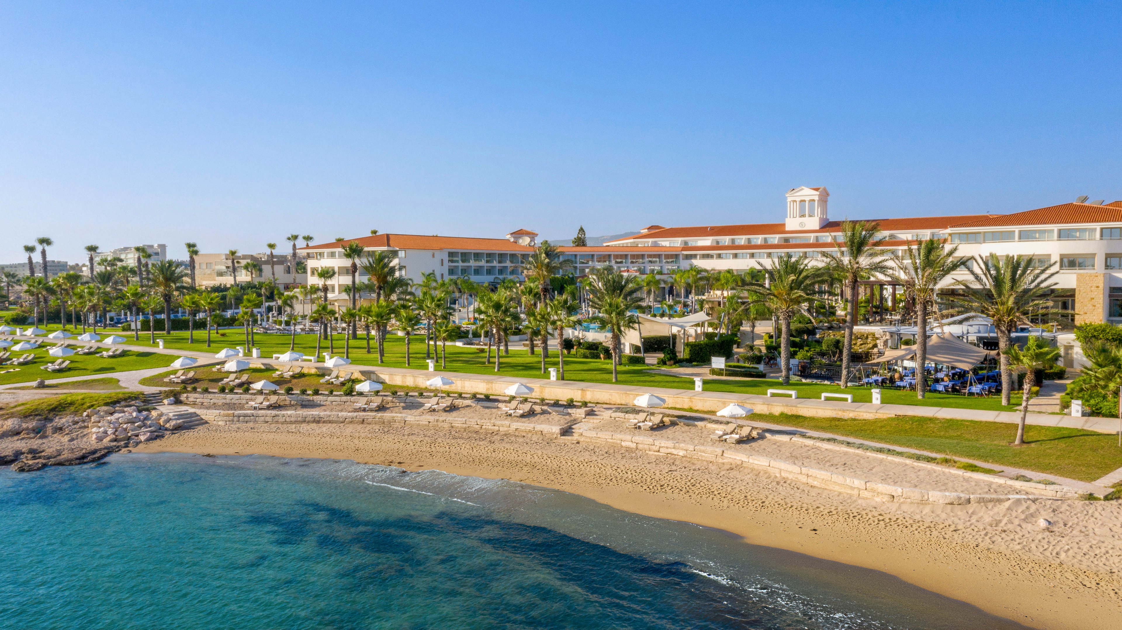 A large beachfront resort hotel stretches along a sandy shoreline beside clear blue water. Palm trees and manicured lawns create a lush, tropical atmosphere around the property. White sun loungers and umbrellas are arranged neatly along the beach and near the hotel grounds. The bright, cloudless sky and calm sea suggest a warm, relaxing vacation destination.