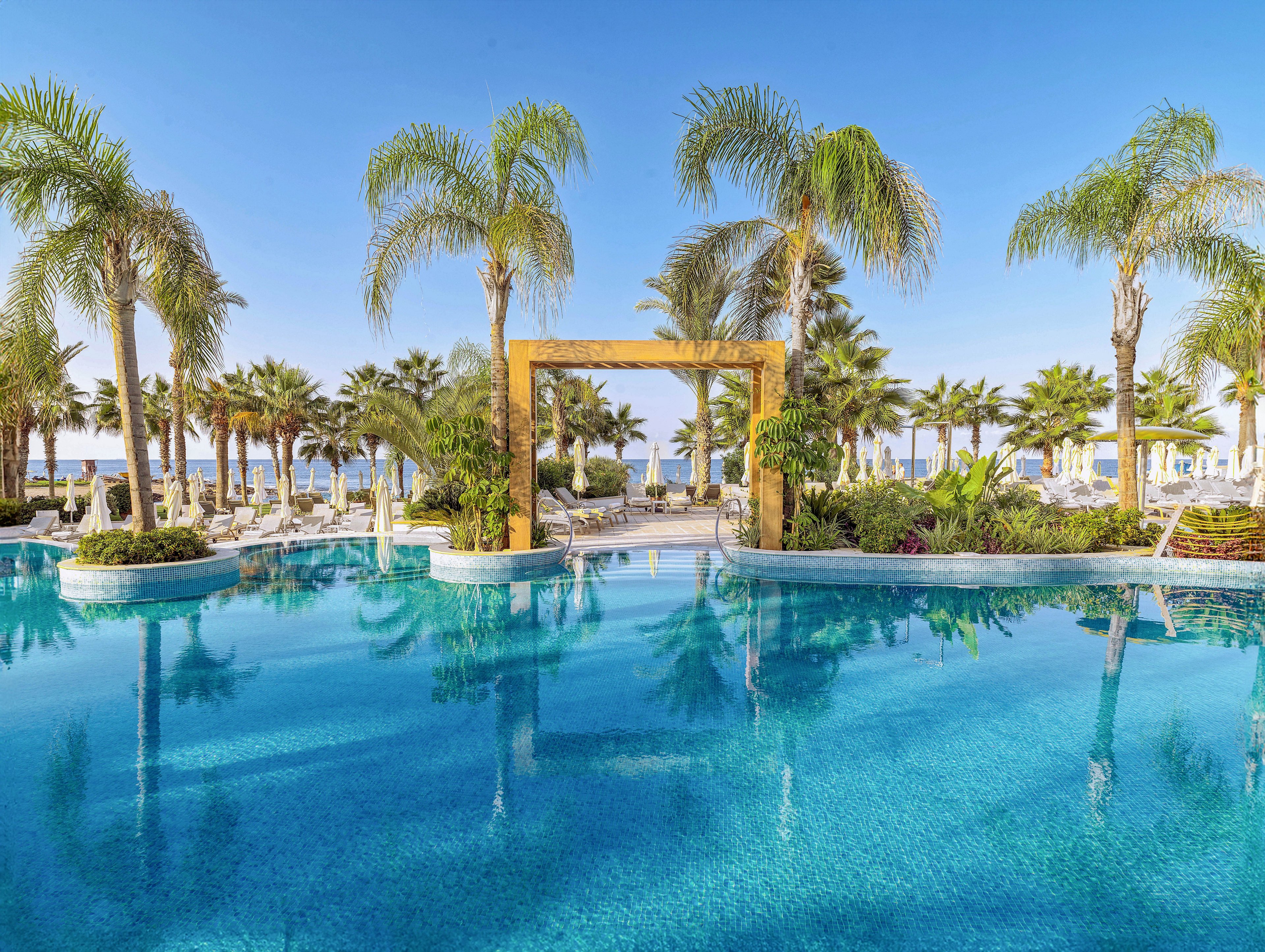 A large, clear blue swimming pool stretches across the foreground of a tropical resort. Tall palm trees and lush greenery surround the pool, framing a decorative wooden archway at its center. Sun loungers and umbrellas line the poolside, with the ocean visible in the distance under a bright blue sky. The scene evokes a serene, luxurious vacation atmosphere in a coastal setting.