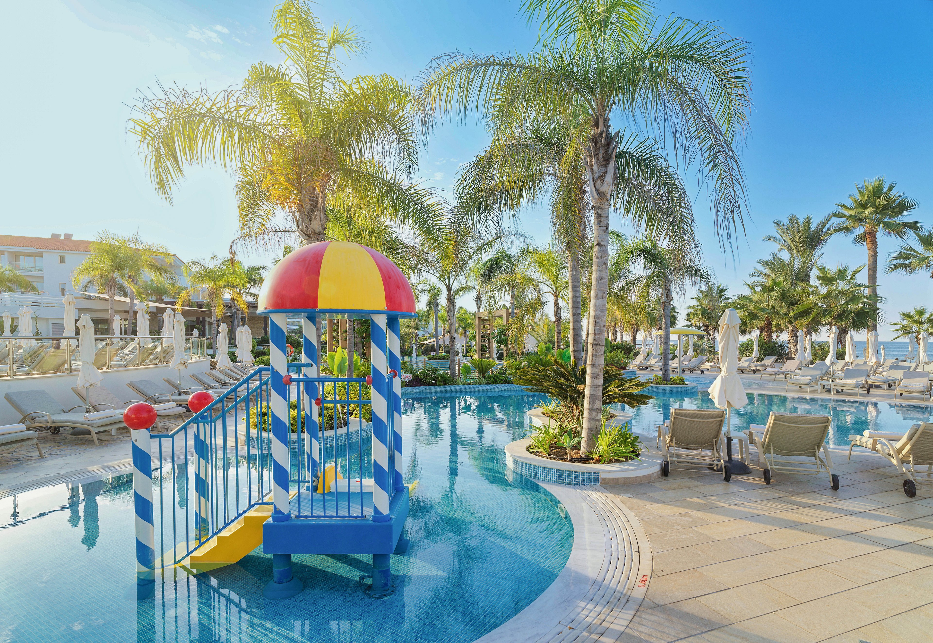 A bright resort swimming pool features a small colorful children's slide structure in the shallow water. Palm trees surround the pool area, creating a tropical vacation atmosphere. Numerous sun loungers and umbrellas line the pool deck, suggesting a relaxing holiday setting. The clear blue sky and calm water emphasize a warm, sunny day ideal for family leisure.