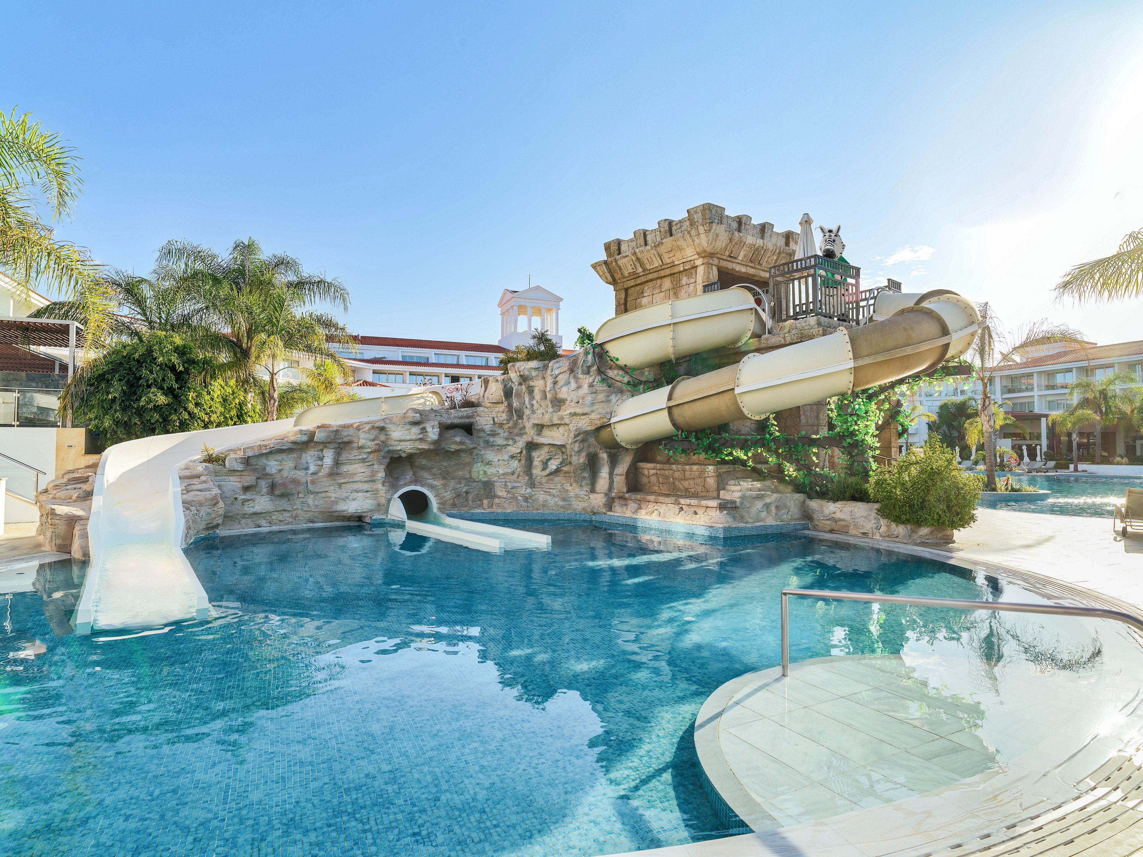 A resort-style swimming pool features a rocky structure with multiple water slides emptying into clear blue water. A young child in a swimsuit slides down a small white slide into the pool, while another child wearing a sun hat and floatation device stands in the shallow area. Palm trees and resort buildings surround the pool, creating a tropical vacation atmosphere. The bright sunlight and clear sky give the scene a warm, cheerful mood.