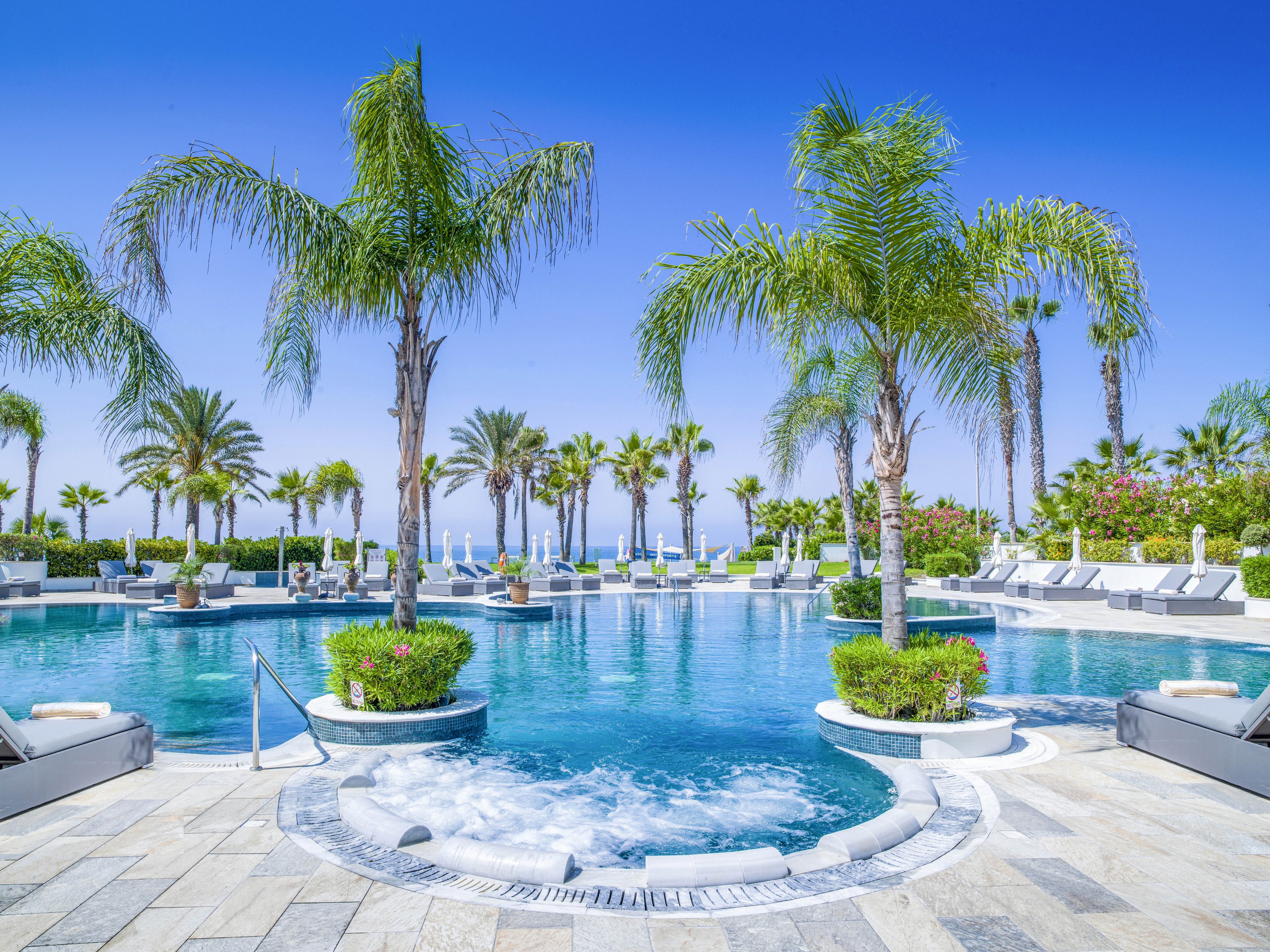 A luxurious resort swimming pool is surrounded by tall palm trees under a clear blue sky. Sun loungers and umbrellas line the poolside, suggesting a relaxing vacation setting. The foreground features a circular spa or hot tub area with bubbling water leading into the larger pool. The scene conveys a bright, sunny, and tranquil tropical atmosphere near the coast.
