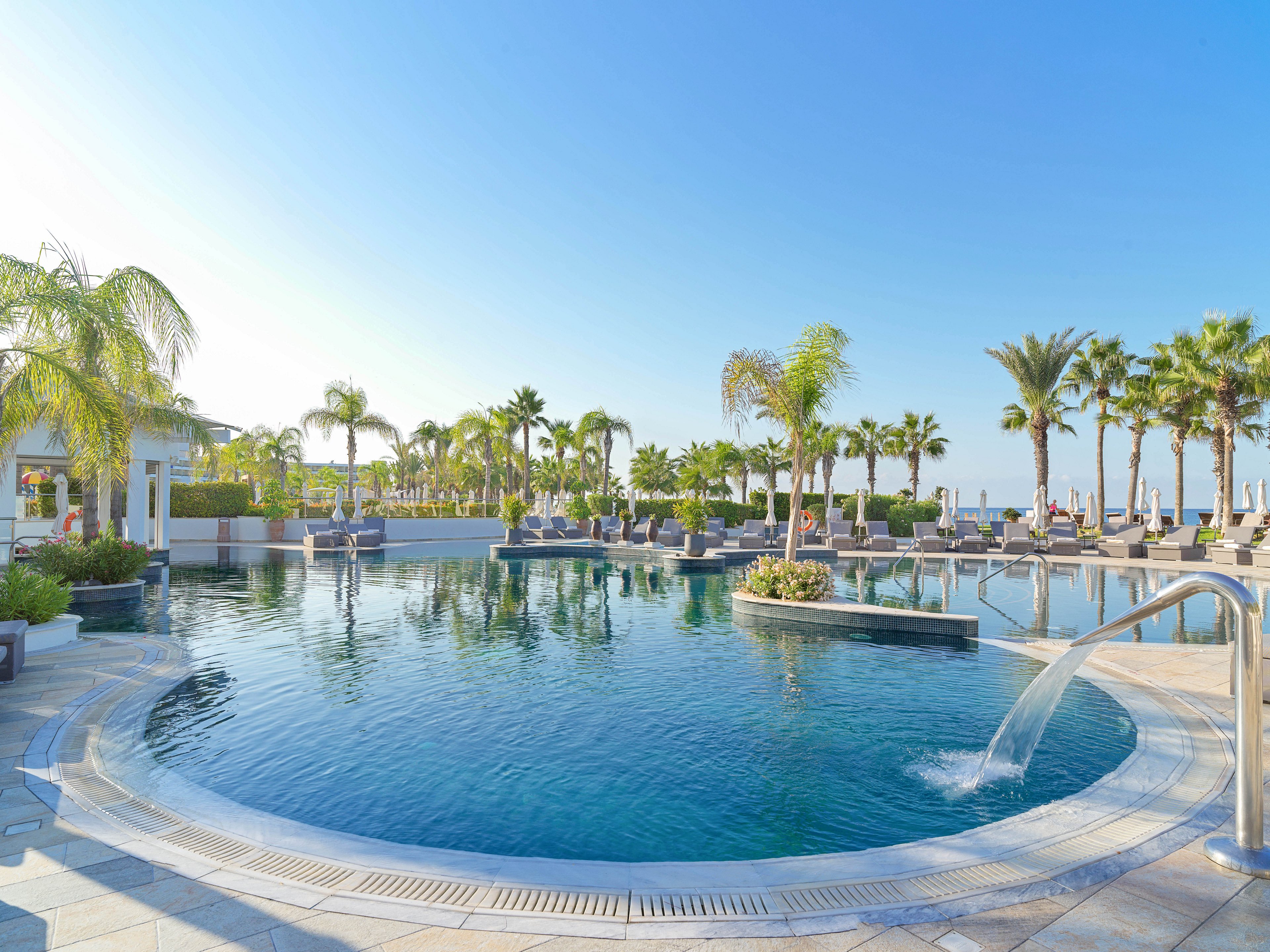 A large circular infinity pool is surrounded by palm trees at a luxury seaside resort. Clear blue water reflects the bright sky, with fountains and water features adding movement to the scene. Sun loungers and umbrellas line the poolside terraces, suggesting a relaxing vacation atmosphere. The overall setting feels tropical and tranquil, ideal for leisure and sunbathing.