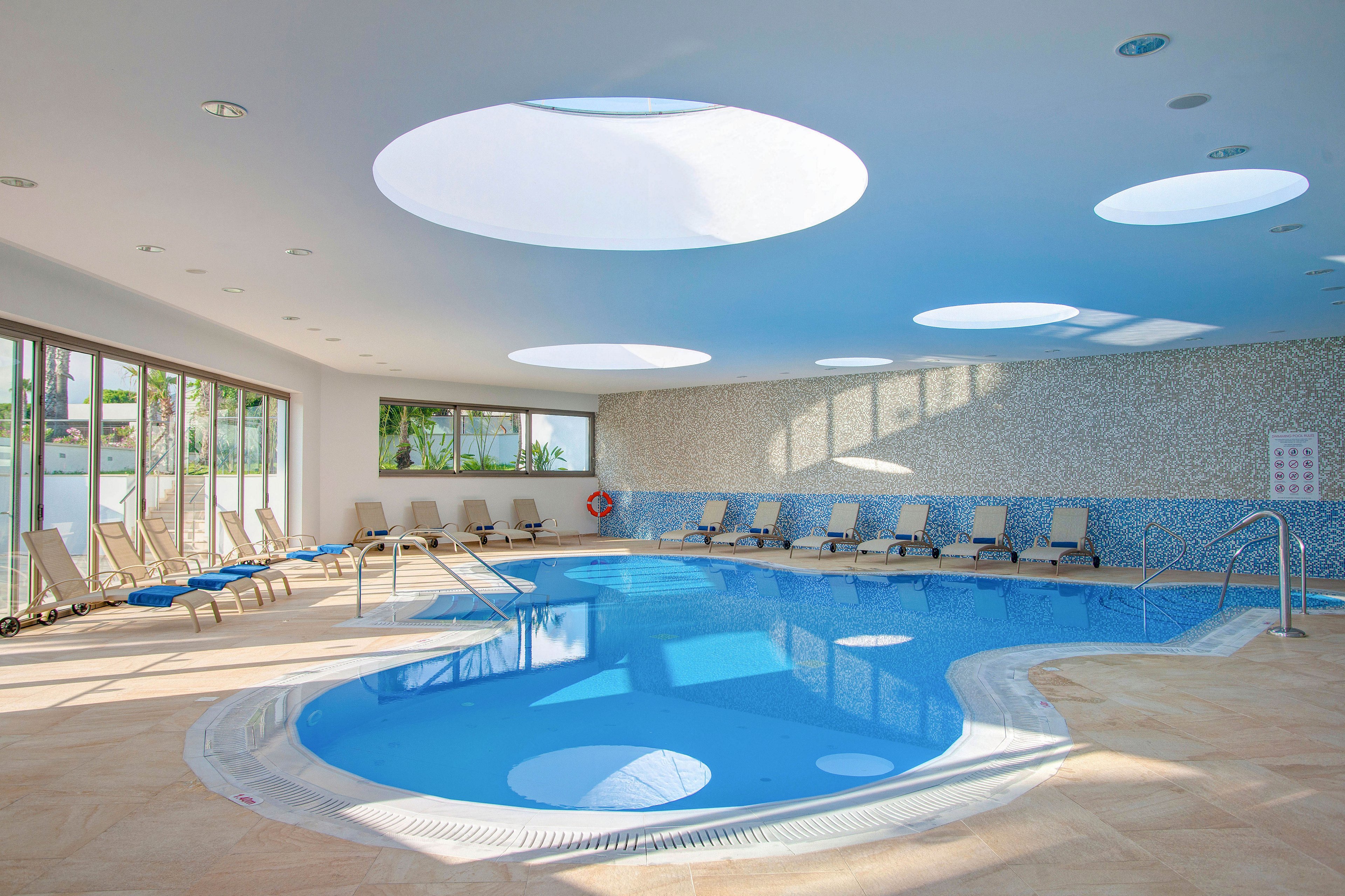 Spacious indoor swimming pool area with clear blue water and modern circular skylights in the ceiling. Numerous beige lounge chairs line the poolside, suggesting a relaxing hotel or resort environment. Large windows on one side let in natural light and offer views of greenery outside. The clean tiled floor and calm, empty space create a serene, spa-like atmosphere.