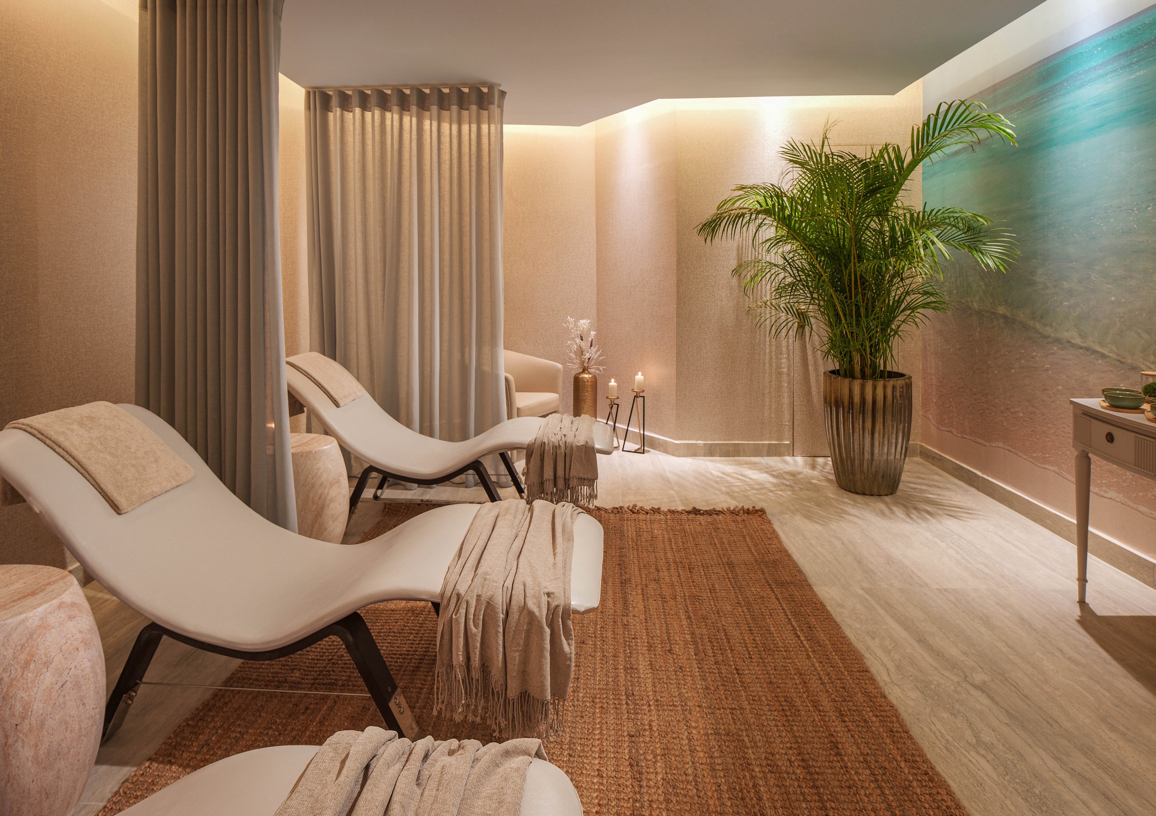 A serene spa relaxation room features two modern reclining loungers draped with soft beige towels. The space is warmly lit, with neutral walls, a woven rug, and floor-length curtains creating a cozy, private atmosphere. A large potted plant and decorative vases add natural and organic elements to the scene. The overall mood is calm, inviting, and designed for rest and wellness.