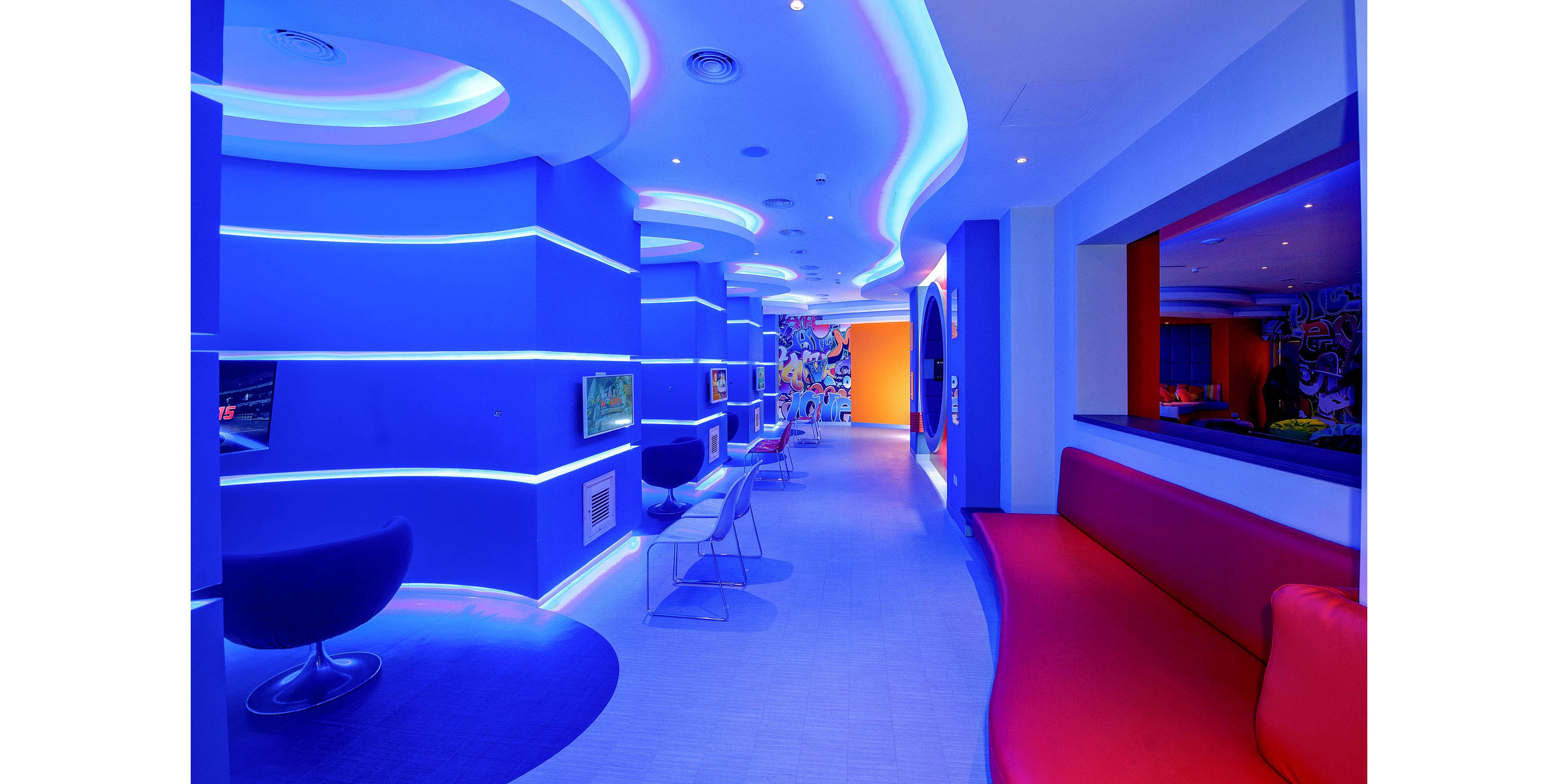 A modern lounge interior is illuminated with intense blue neon lighting and curved ceiling details. Sleek chairs and small tables line the corridor, creating a contemporary waiting or social area. A long red bench contrasts with the blue walls, while recessed screens and wall cutouts add visual interest. The space feels futuristic, colorful, and dynamic, with no people present.