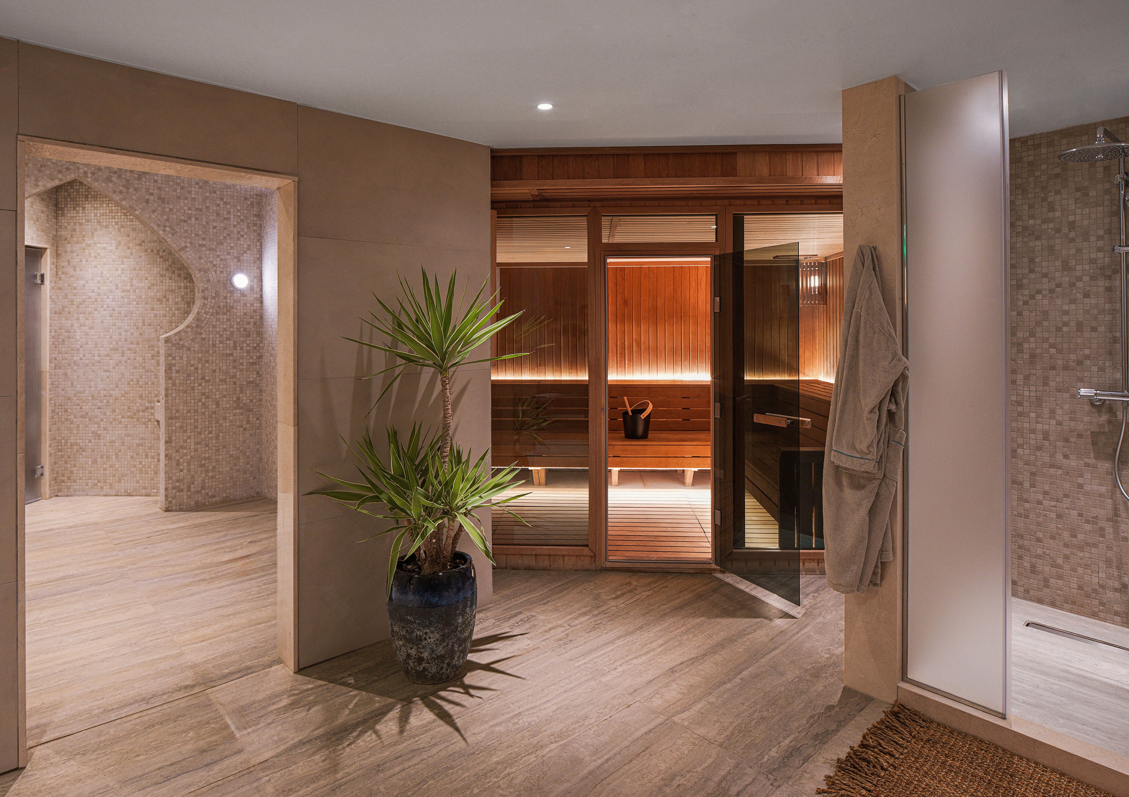 A contemporary spa interior features a warm wooden sauna room with soft indirect lighting. The sauna is viewed through glass doors from a spacious tiled relaxation area. A potted plant, hanging robe, and textured walls add to the serene atmosphere. Neutral tones and clean lines create a calm, minimalist wellness setting.