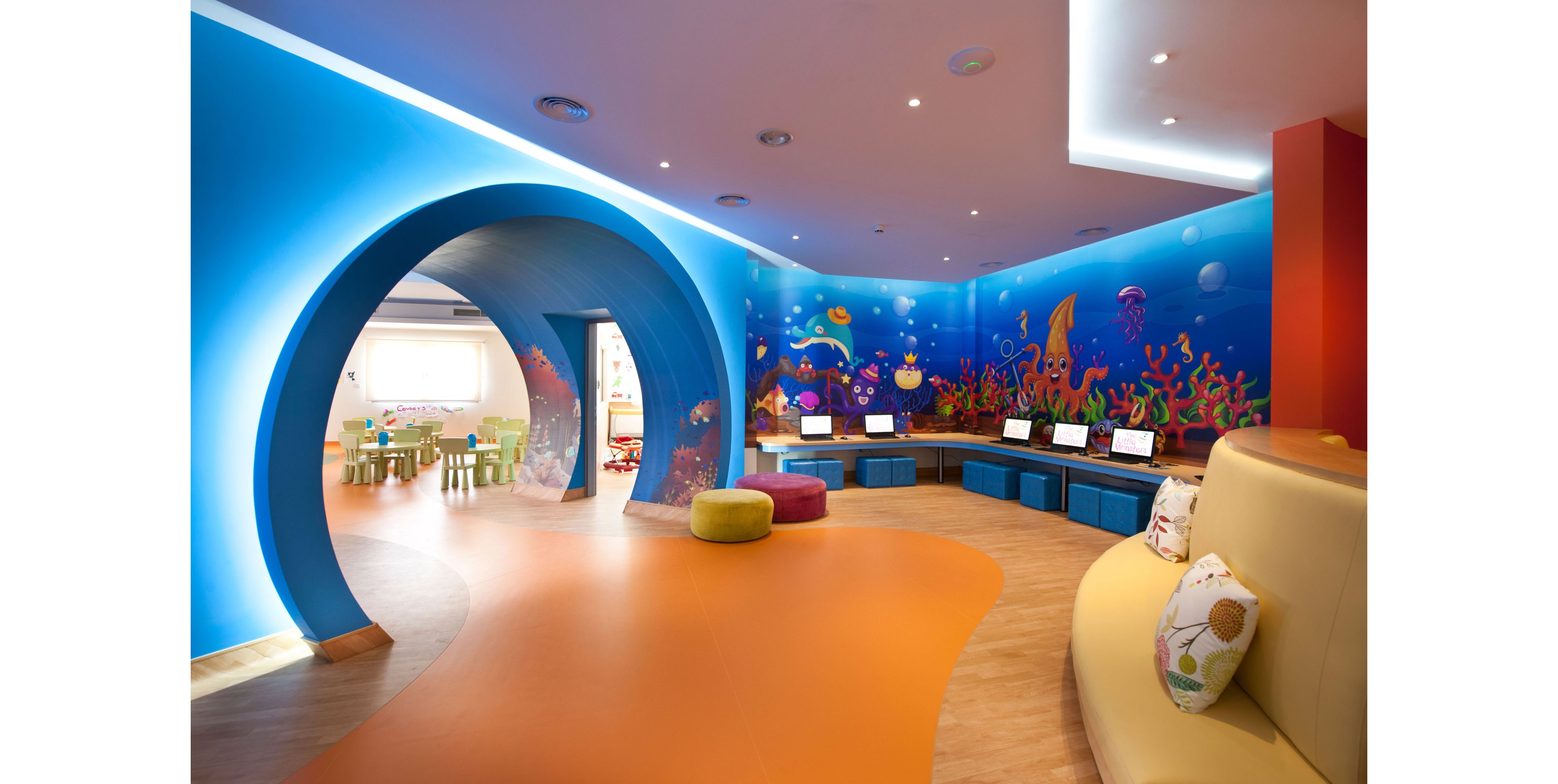 A brightly colored children’s playroom features an underwater-themed mural with fish, coral, and sea creatures covering the curved blue walls. An arched tunnel leads to a smaller area with child-sized tables and chairs in the background. In the foreground, cushioned seating and poufs sit on an orange and wood-tone floor, while a row of computers is arranged along the wall. Recessed ceiling lights and soft indirect lighting create a cheerful, welcoming atmosphere.