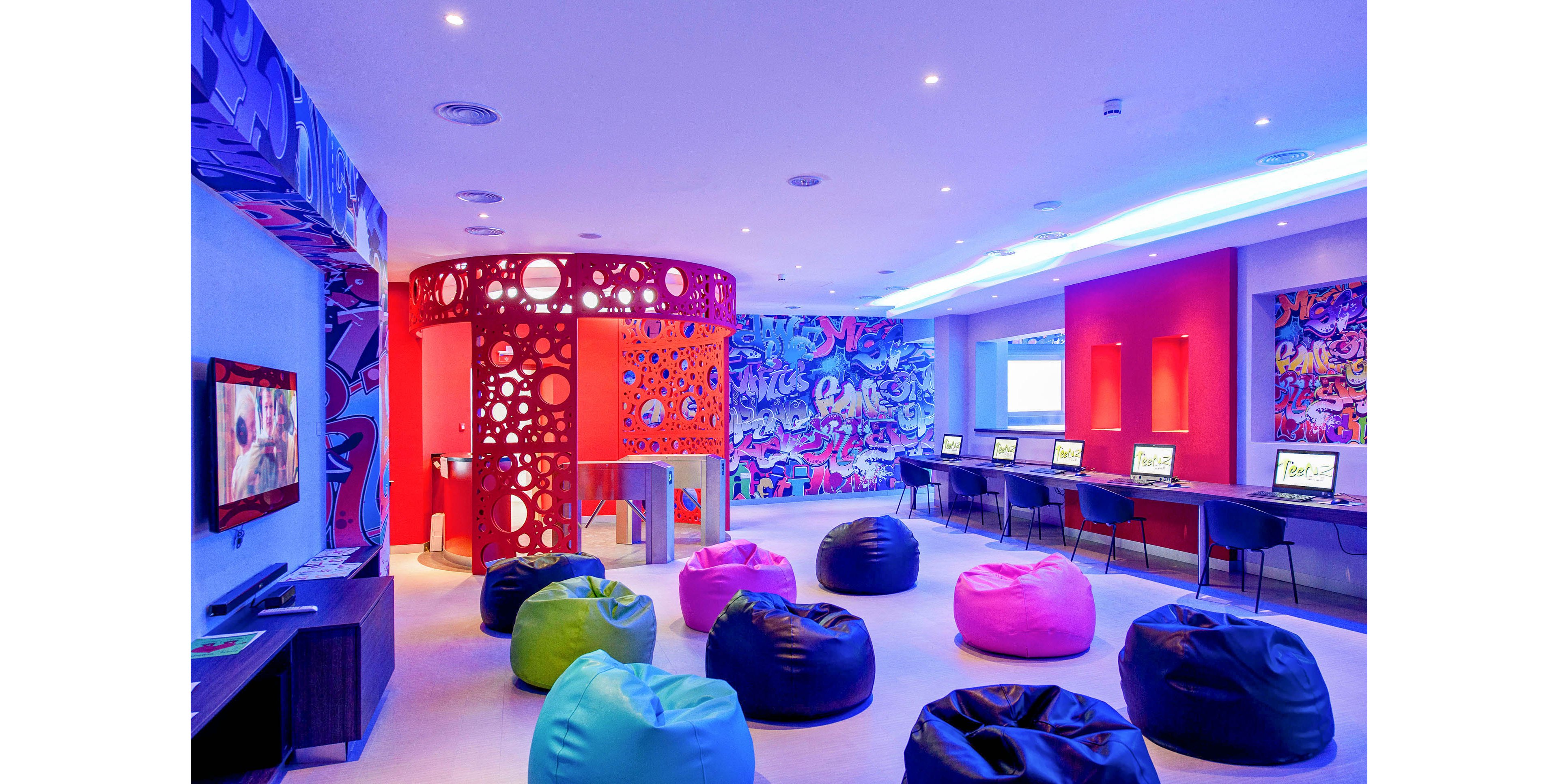 A vibrant contemporary lounge features numerous colorful bean bag chairs arranged across a spacious room. Along one wall, a row of computers sits on a long desk, creating a casual workspace or internet area. A striking red circular structure with cut-out patterns stands near the center, complemented by bold graffiti-style wall art in bright blues, pinks, and purples. Recessed ceiling lights and blue accent lighting give the space a futuristic, playful atmosphere.