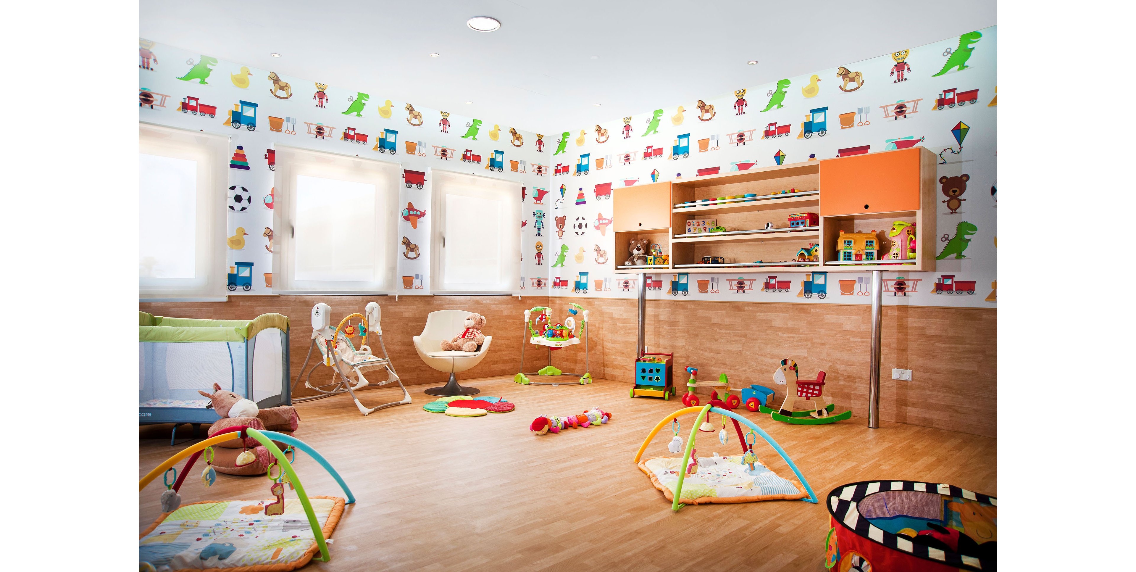 Colorful daycare playroom with various toys and play mats arranged on a wooden floor. The room features large windows with soft light filtering through white blinds. Walls are decorated with playful animal and vehicle illustrations, creating a cheerful atmosphere. Shelves hold stuffed animals, blocks, and other educational toys, while baby bouncers and activity gyms are placed around the room.