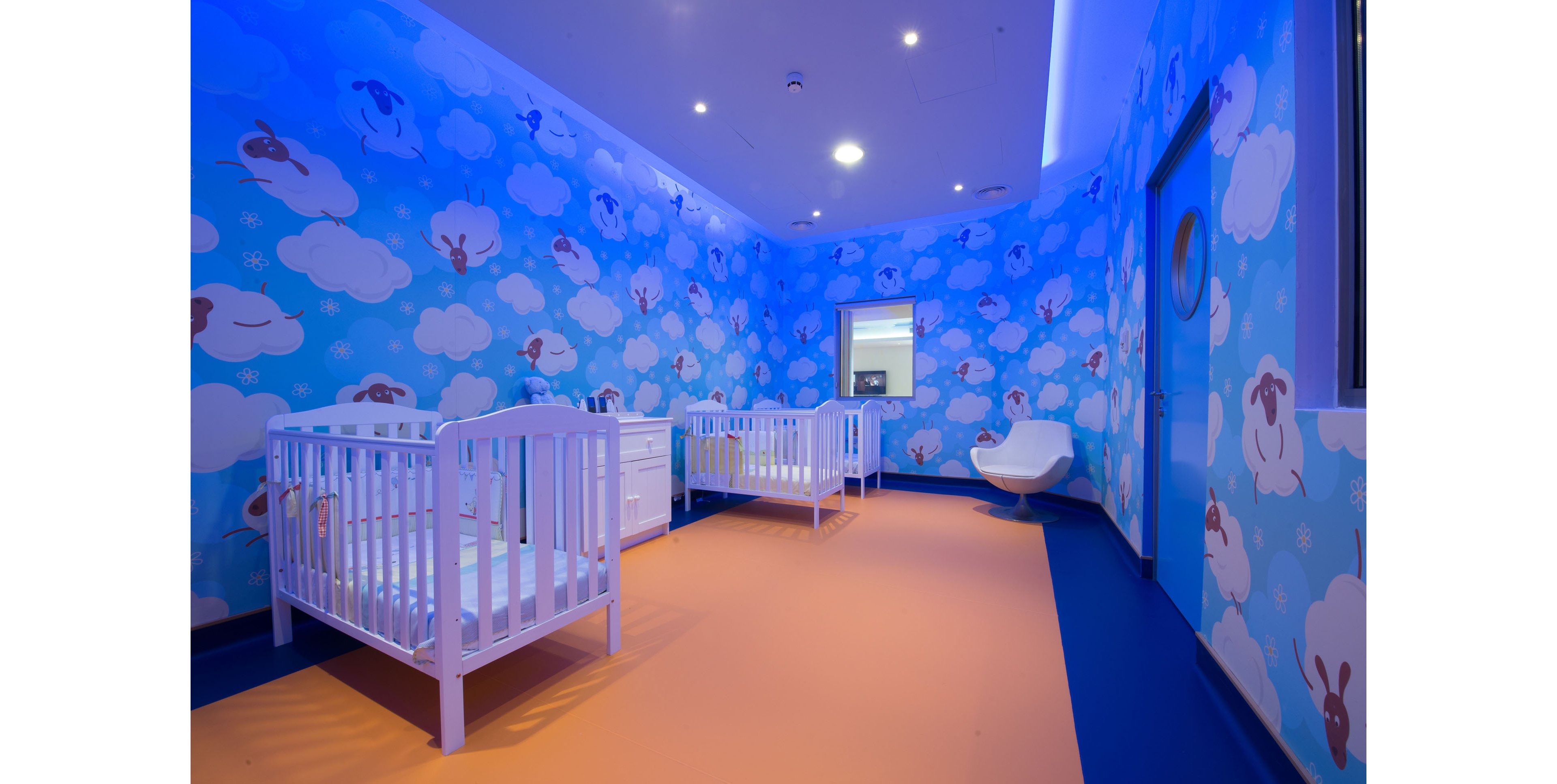 A brightly lit nursery room features two white baby cribs placed against a whimsical blue wall. The walls are decorated with cartoon clouds and sheep, creating a playful and dreamy atmosphere. The floor combines warm orange and blue tones, adding contrast to the cool blue walls. A modern white chair and a small dresser complete the minimal, child-friendly interior.