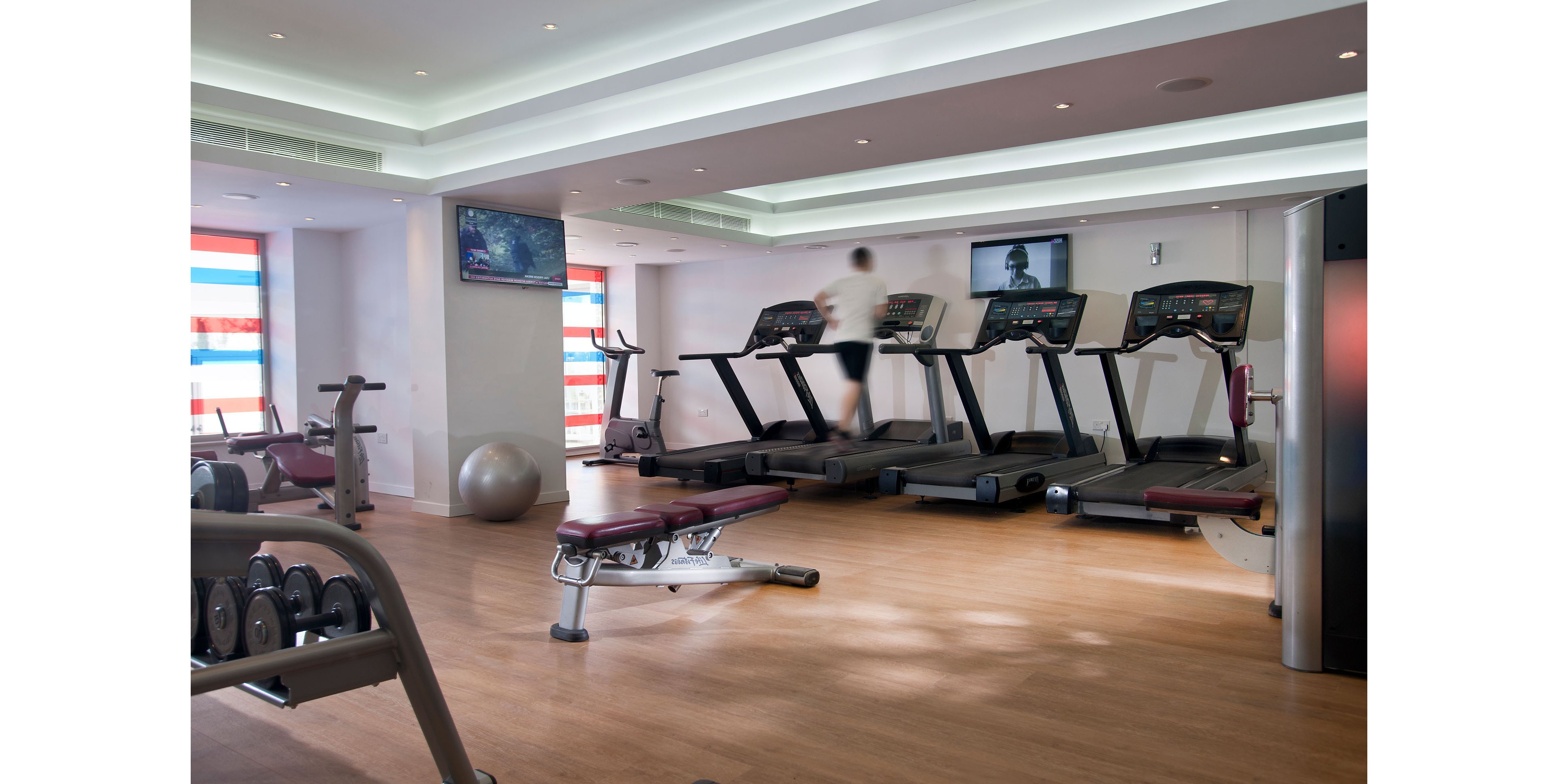 A man sits on a bench in a bright fitness center, lifting a dumbbell with one arm. Several treadmills line the back wall, with another person running on one of them. The gym features wooden flooring, white walls, and recessed ceiling lights, creating a clean and modern atmosphere. Free weights and benches are visible in the foreground, emphasizing strength and cardio training equipment.