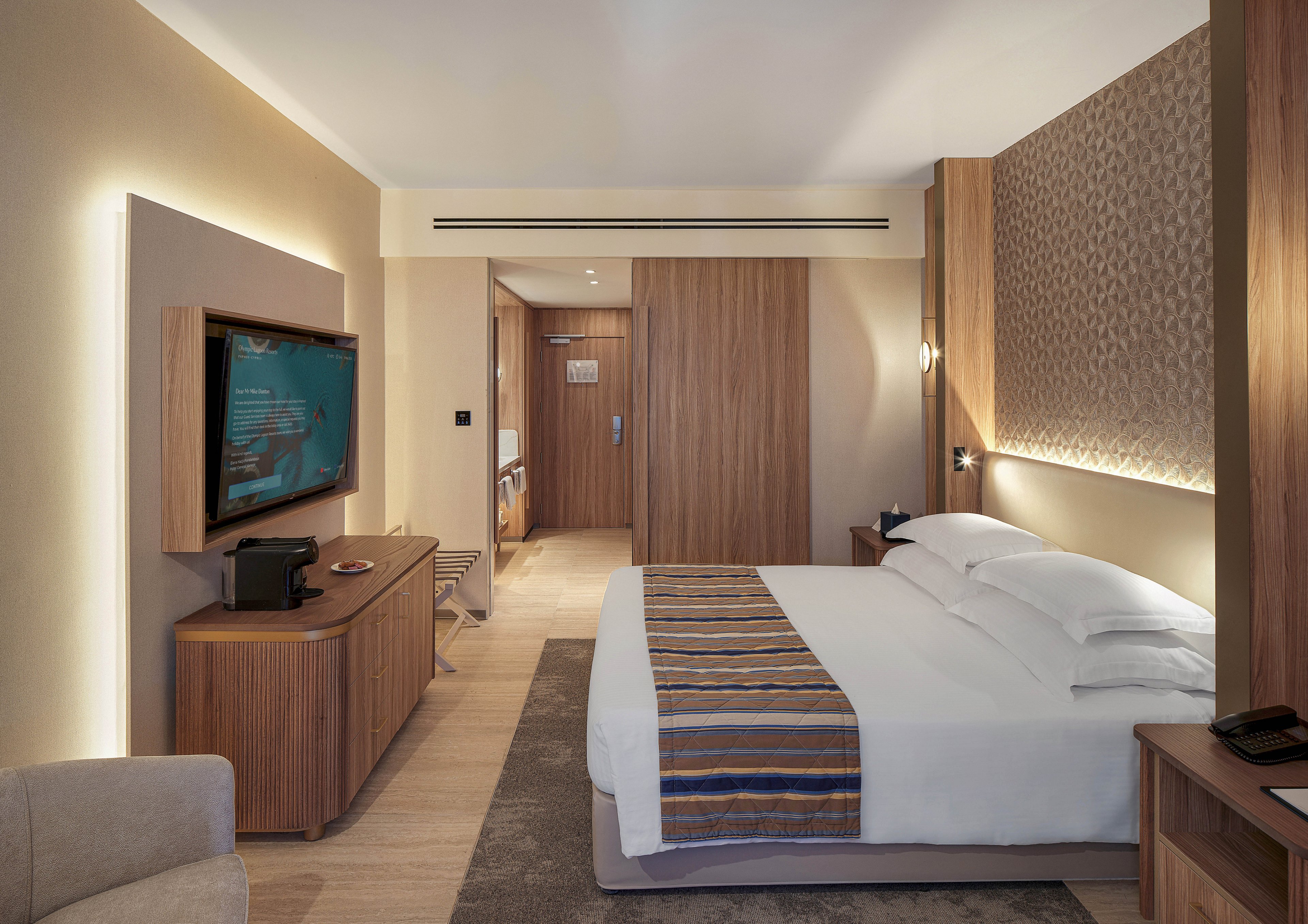 Contemporary hotel room featuring a large neatly made bed with white linens and a striped runner. The space includes warm wooden furniture, a wall-mounted television, and a small coffee station on a sideboard. Soft ambient lighting highlights the textured accent wall behind the bed and the clean lines of the decor. A doorway at the back reveals the entrance corridor and bathroom area, emphasizing a comfortable and inviting stay.