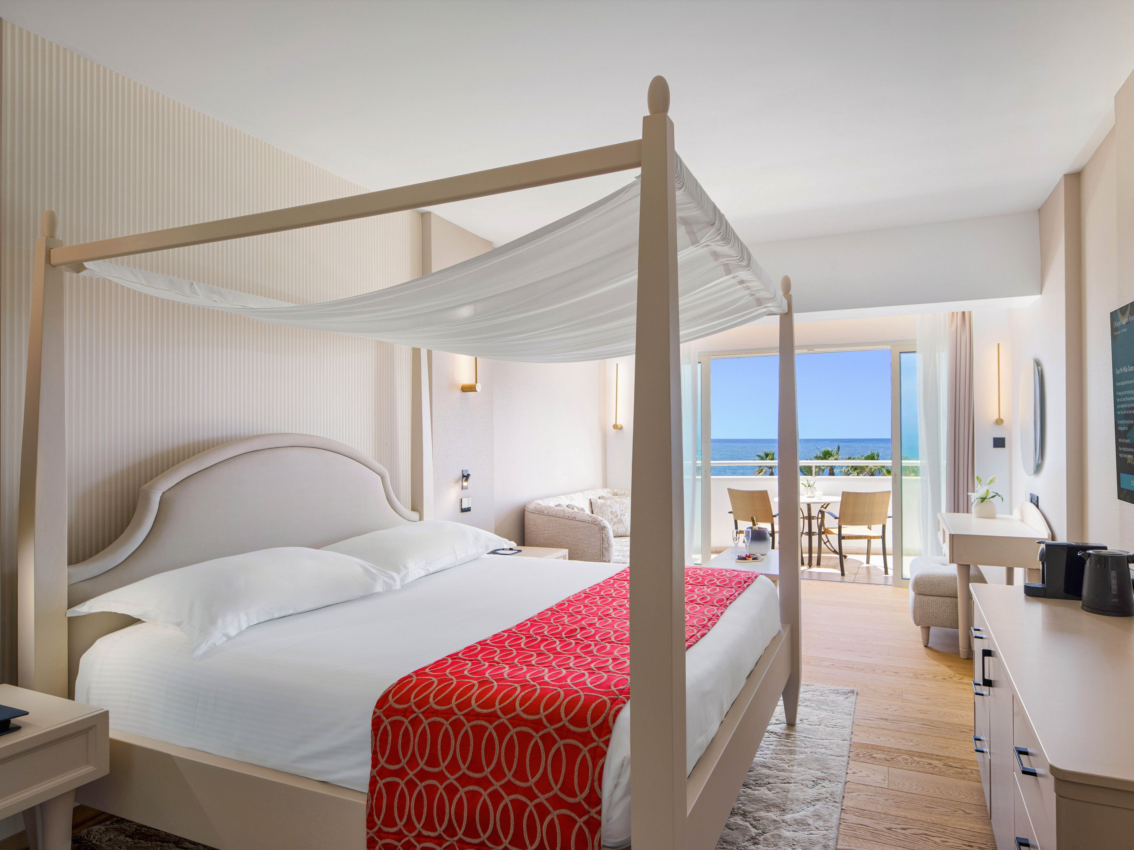 Spacious hotel room featuring a four-poster bed with white linens and a red patterned throw. The interior is decorated in light neutral tones with wooden flooring and modern furnishings. A sitting area and desk lead toward large glass doors opening to a balcony with outdoor seating. Beyond the balcony, a clear view of the sea and blue sky enhances the airy, relaxing atmosphere.