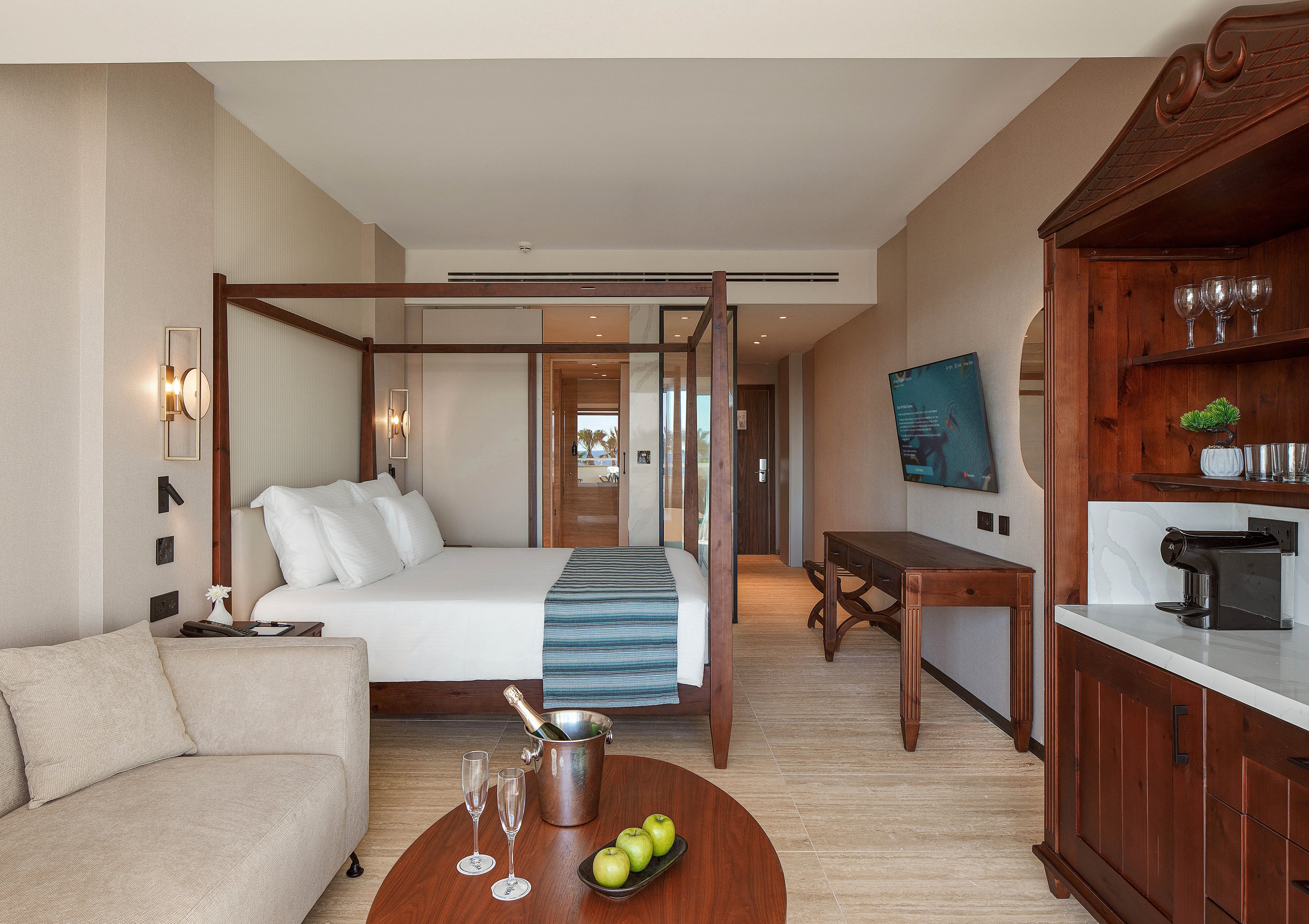 A contemporary hotel room features a four-poster bed with crisp white linens and a teal runner. The space includes a light-colored sofa, a wooden table with green apples and champagne glasses, and a wall-mounted flat-screen TV. A wooden cabinet with a coffee machine and glassware sits beside a white countertop. Neutral walls and flooring, along with warm wood furniture, create a calm and inviting atmosphere.