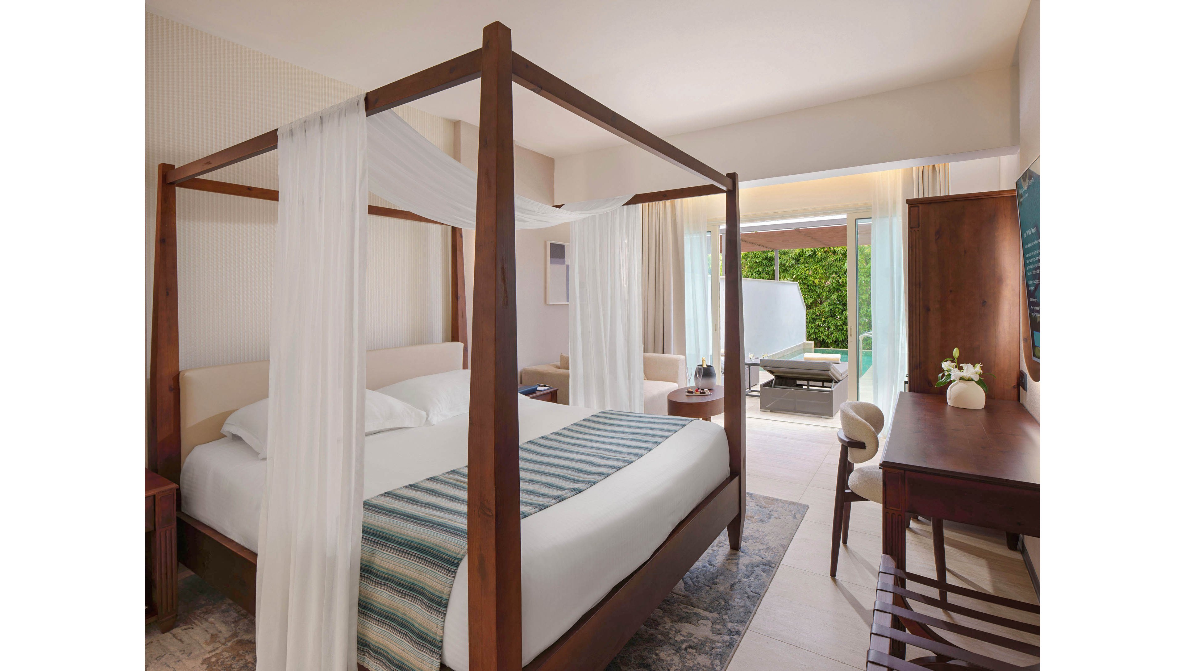 A contemporary hotel bedroom features a wooden four-poster canopy bed with white drapes and striped blue runner. The room includes a wooden desk, chair, and bench, with neutral walls and light flooring creating an airy atmosphere. Large sliding glass doors open to a private terrace with lounge seating and greenery outside. Soft natural light fills the space, emphasizing the clean, minimalist design and relaxing resort feel.