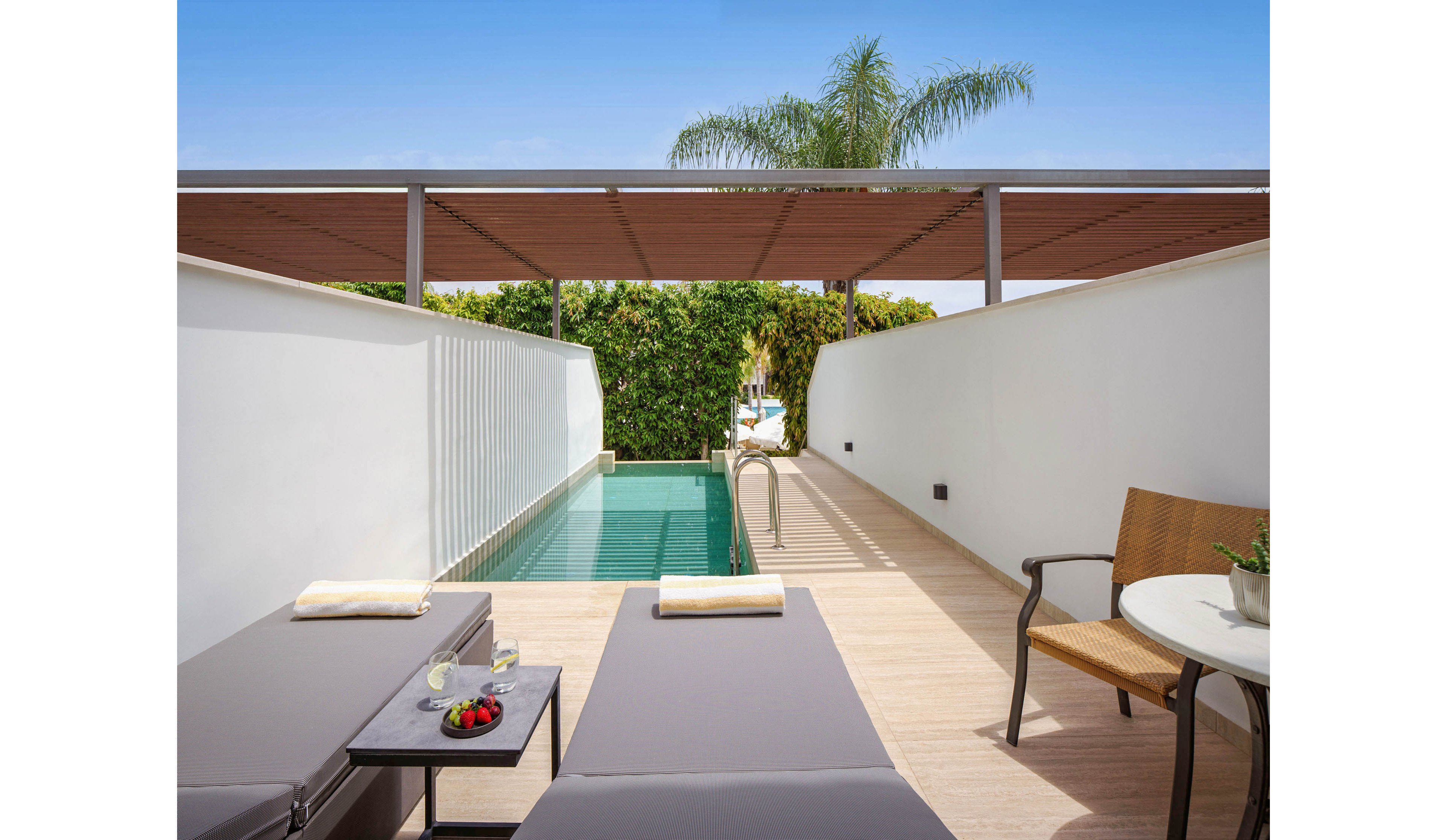 A contemporary private terrace features two cushioned loungers beside a narrow plunge pool. The space is framed by white walls, a wooden deck, and a slatted pergola overhead. A small side table holds drinks and a plate of strawberries, while a chair and round table sit nearby for relaxation. Lush greenery and a palm tree at the far end add a tropical resort atmosphere.