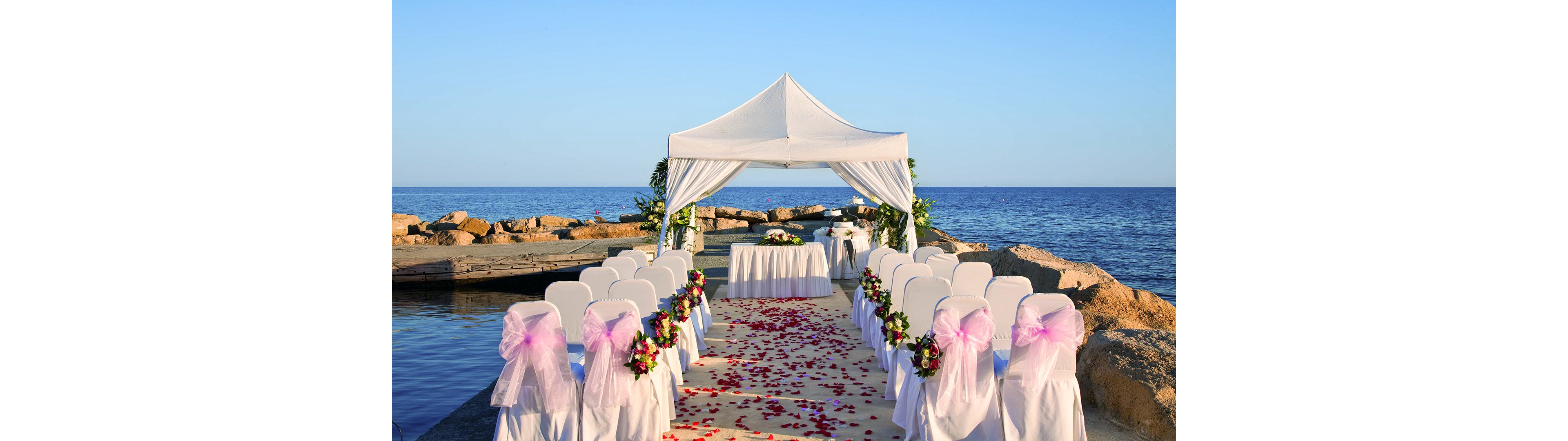An elegant wedding ceremony setup is arranged on a stone pier by the sea under a white canopy. Two rows of white chairs with pink ribbons face a small table at the front, creating a central aisle. The aisle is covered with a light-colored runner scattered with red rose petals. Colorful floral arrangements decorate the chairs and canopy, while calm blue water and clear sky form a serene backdrop.