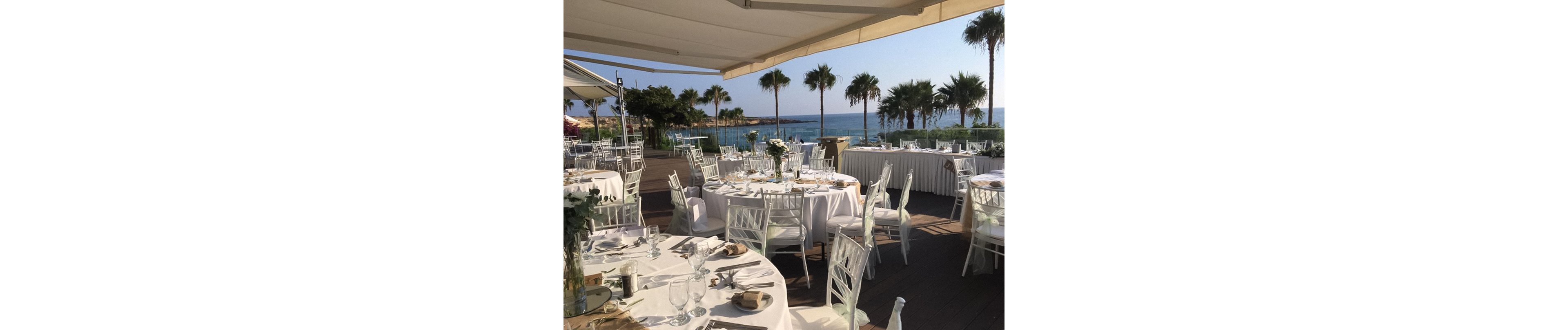 An elegant outdoor reception is set up on a wooden deck overlooking the sea. Round tables with white linens, glassware, and floral centerpieces are arranged beneath large awnings. White chiavari chairs surround the tables, creating a refined and cohesive look. Palm trees, calm blue water, and clear skies provide a serene coastal backdrop. The scene suggests a formal wedding or special event in a warm, seaside location.