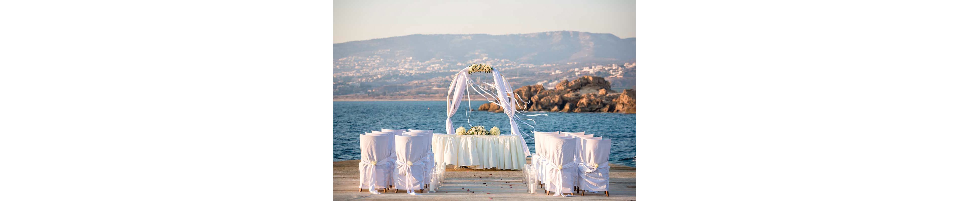 An elegant wedding ceremony setup is arranged on a pier overlooking a calm sea. White chairs wrapped in fabric and bows face a round floral arch draped with flowing ribbons. A table covered with a white cloth and floral arrangements stands beneath the arch. Rocky coastline and distant hills create a scenic backdrop in the warm evening light.