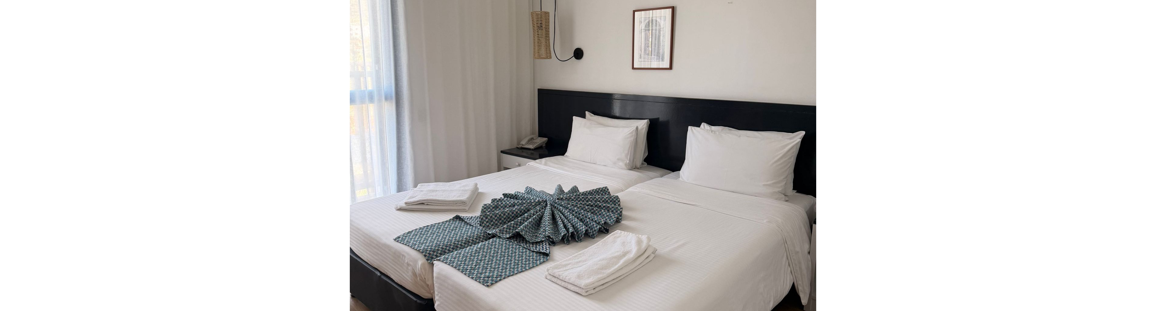 A contemporary hotel room features two neatly made twin beds with crisp white linens and folded towels arranged on top. The room is well-lit with natural light streaming through sheer curtains, and a wall-mounted air conditioner is visible above the beds. Minimalist decor includes a framed picture and a small bedside table, creating a clean and inviting atmosphere.
