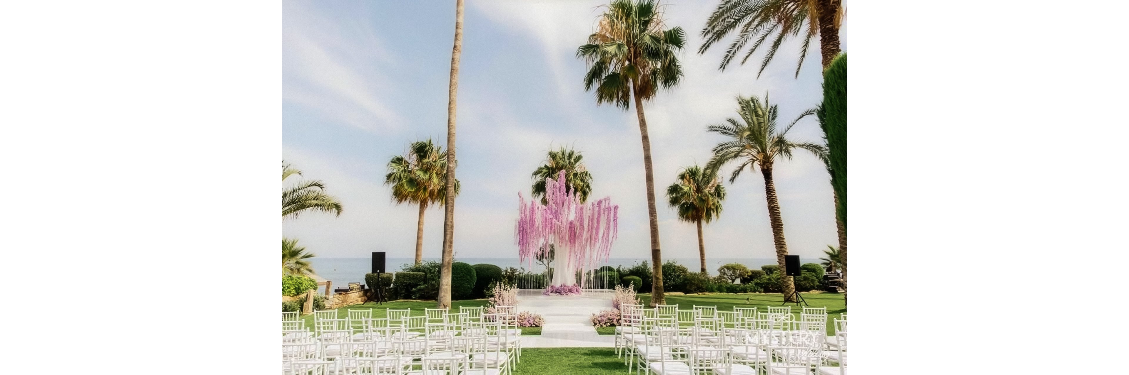 An elegant outdoor wedding ceremony setup is arranged on a manicured lawn overlooking the sea. White chairs are lined up in rows facing a central aisle that leads to a floral altar. The altar features cascading pink and lavender flowers framed by tall palm trees and lush greenery. The bright daylight and soft pastel colors create a romantic, tropical atmosphere.