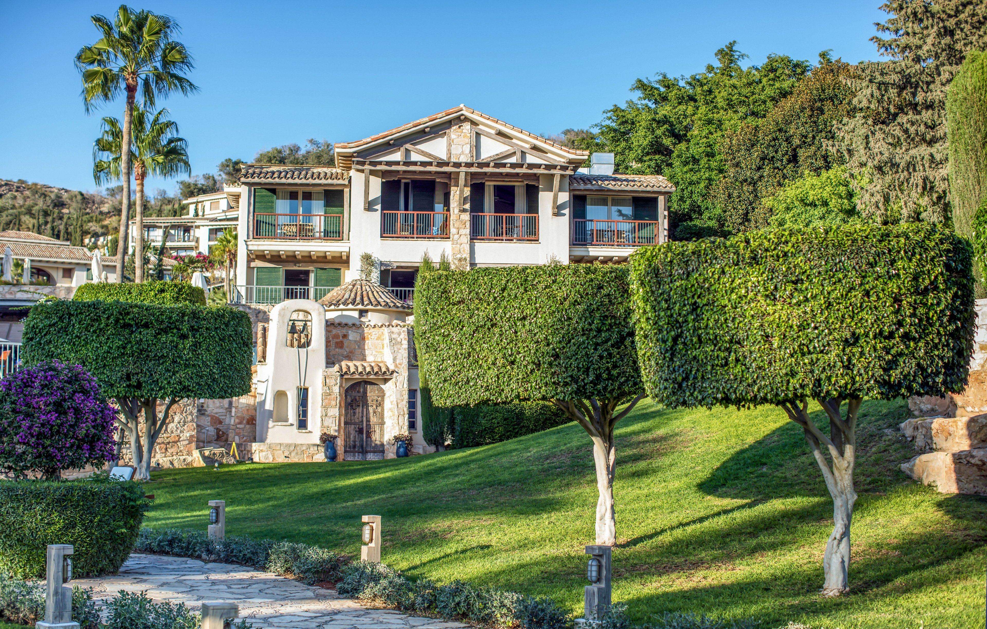 A multi-story Mediterranean-style villa stands prominently in a lush landscaped garden. The foreground features neatly trimmed geometric trees and a stone pathway leading toward the house. Palm trees and surrounding greenery frame the building, suggesting a warm, coastal climate. Sunlight highlights the white walls, terracotta roof tiles, and balconies, creating a bright and inviting atmosphere.