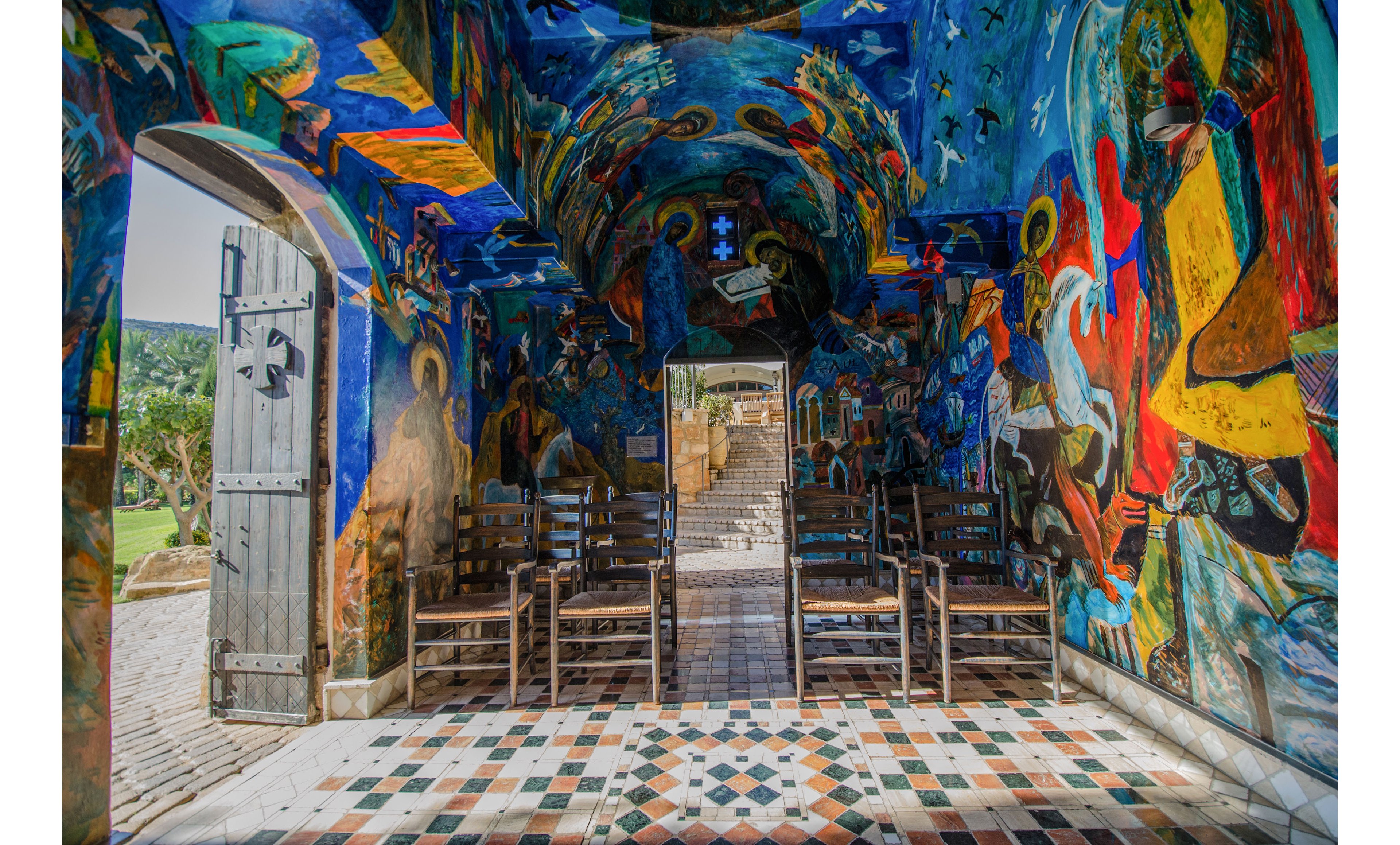 A small chapel interior is richly decorated with colorful frescoes covering the walls and arched ceiling. Wooden chairs are arranged in rows facing a central aisle that leads toward stone steps and an open doorway. The open door on the left reveals a glimpse of the surrounding landscape and bright daylight outside. Intricate geometric tiles in various colors form a patterned floor that guides the viewer’s eye into the vivid, spiritual space.