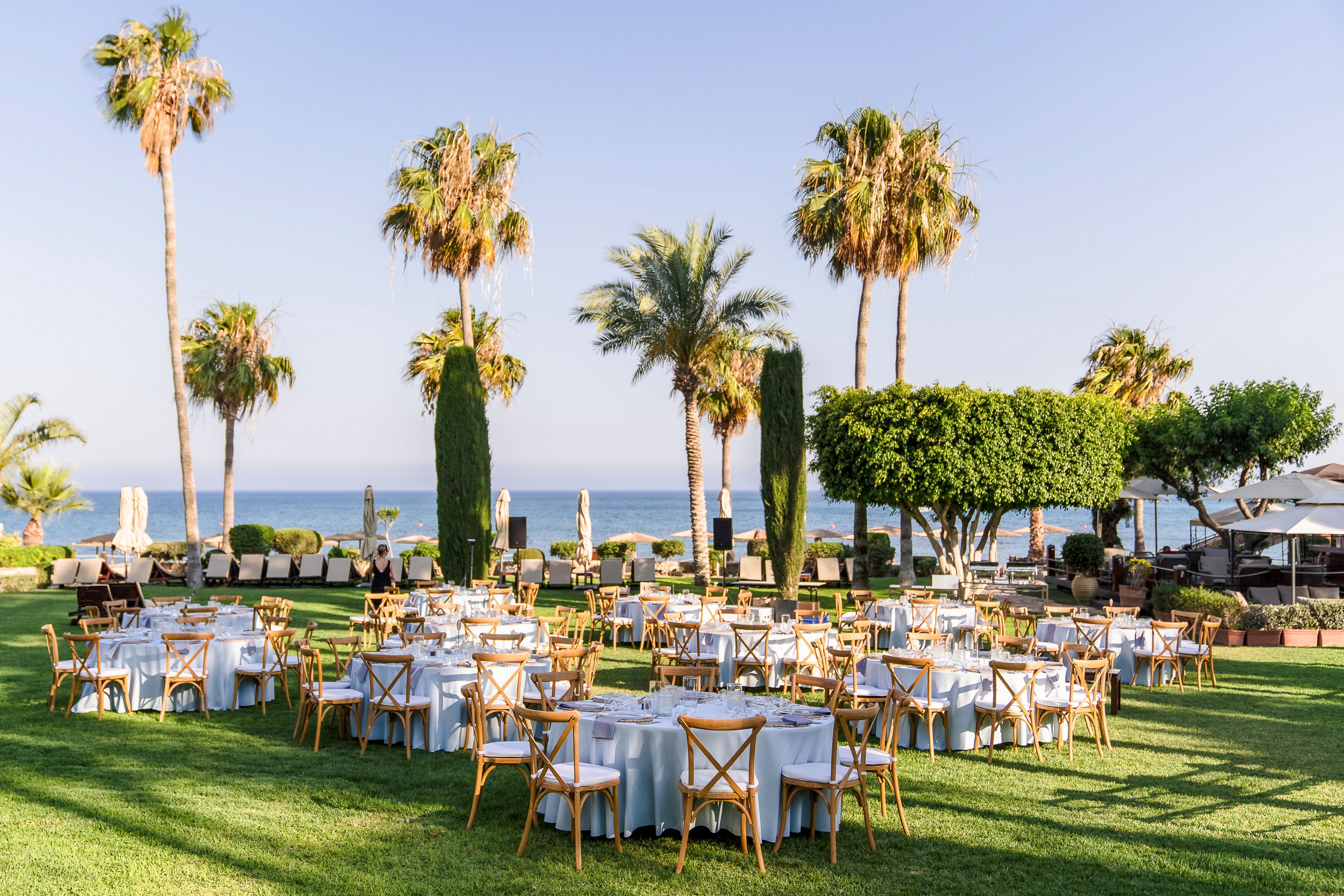 An elegant outdoor event setup features multiple round tables with white tablecloths arranged on a manicured lawn. Wooden chairs surround each table, creating a formal yet relaxed atmosphere. Tall palm trees and trimmed green shrubs frame the scene, with a clear view of the ocean and blue sky in the background. The bright daylight and soft colors suggest a warm, sunny coastal gathering.