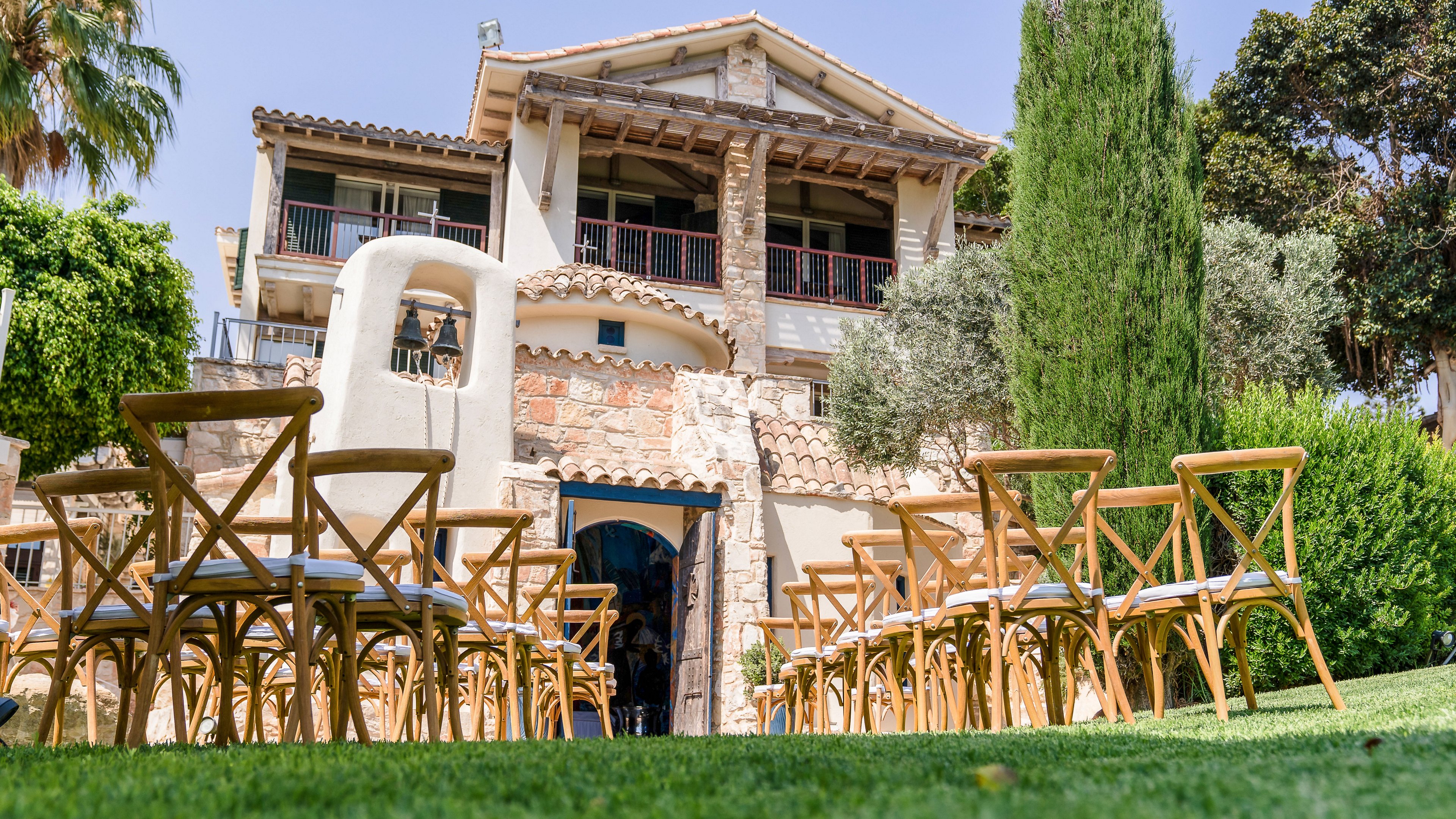 Rows of wooden chairs are arranged on a green lawn facing a rustic stone villa, suggesting preparation for an outdoor ceremony or event. The house features balconies, wooden beams, and a mix of stone and stucco details, framed by lush trees and plants. Bright daylight and clear skies create a warm, inviting atmosphere. The perspective from the grass level emphasizes the aisle leading toward the building entrance.